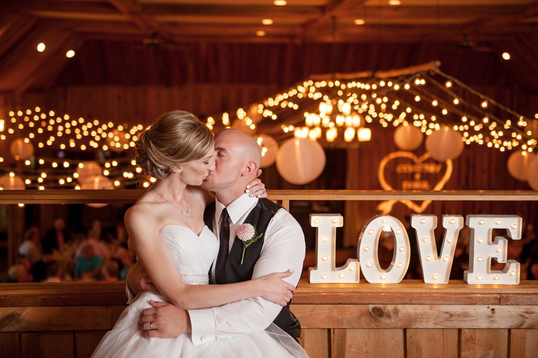 Bride and groom kissing at a warmly lit wedding reception, with a large “LOVE” sign behind them
