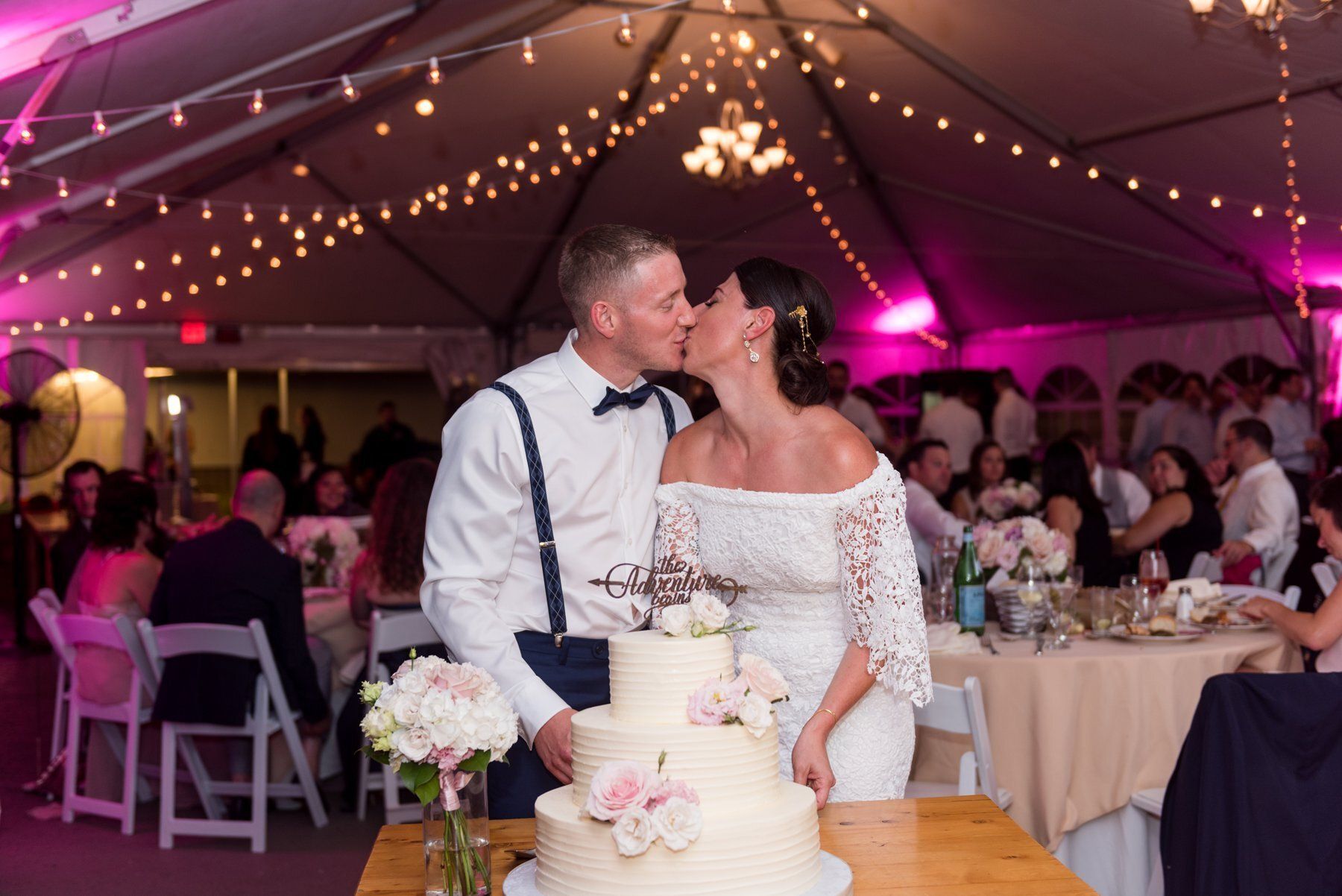 Couple kissing beside a wedding cake under pink-lit tent with string lights
