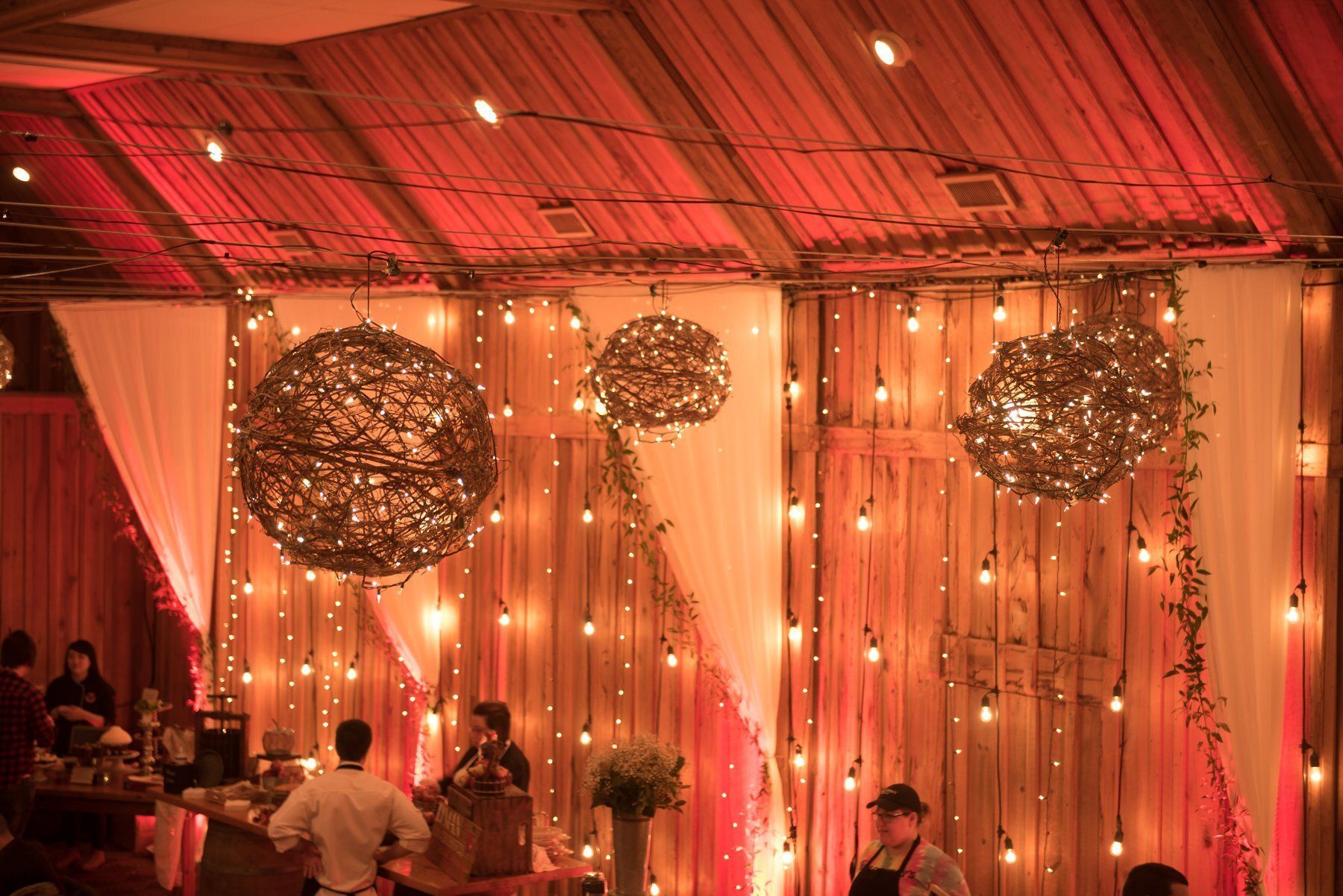 Decorated event hall with warm pink lighting, hanging globe lights, draped curtains, and seated guests