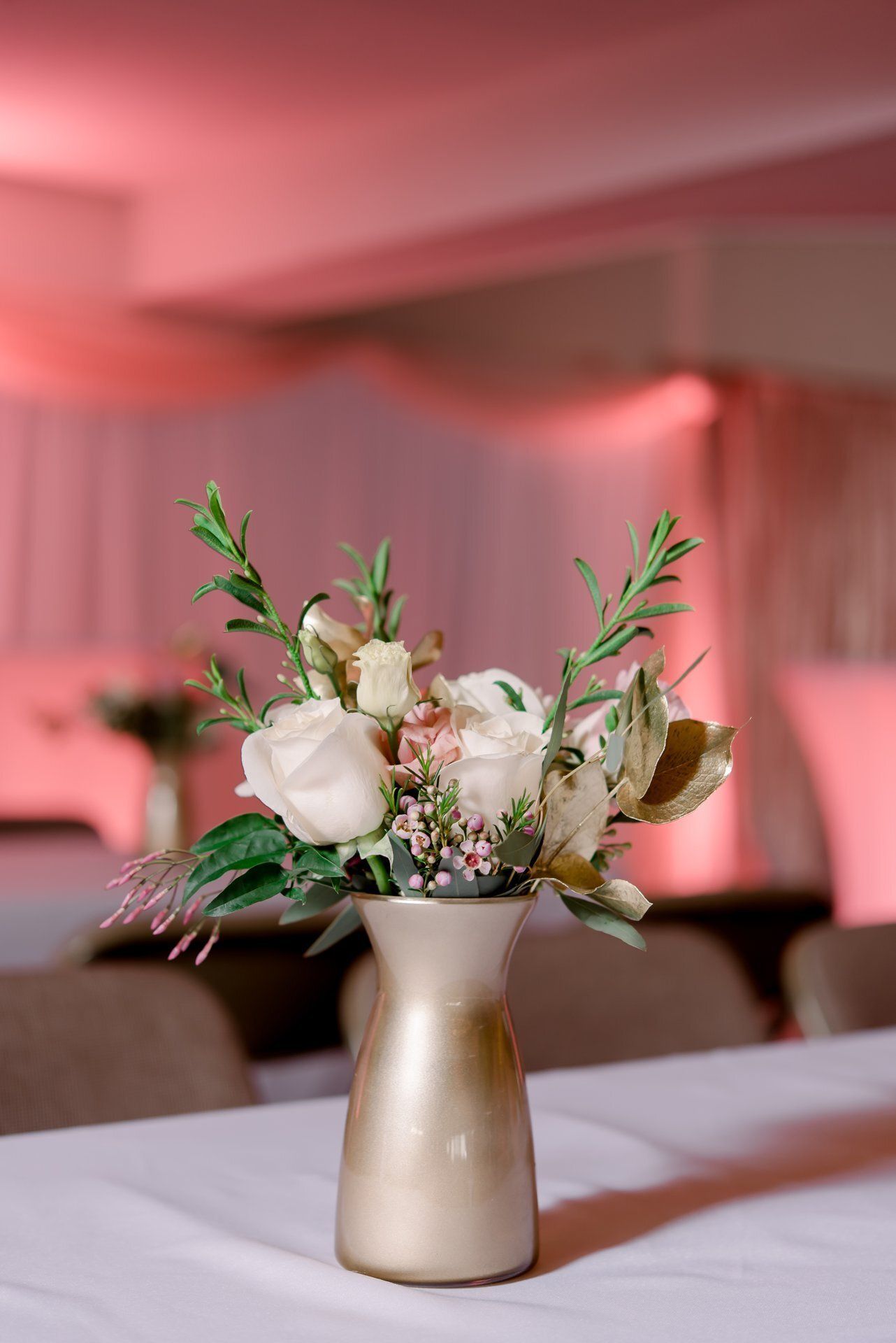 Small floral bouquet in a gold vase on a table, with a pink-lit event backdrop behind it