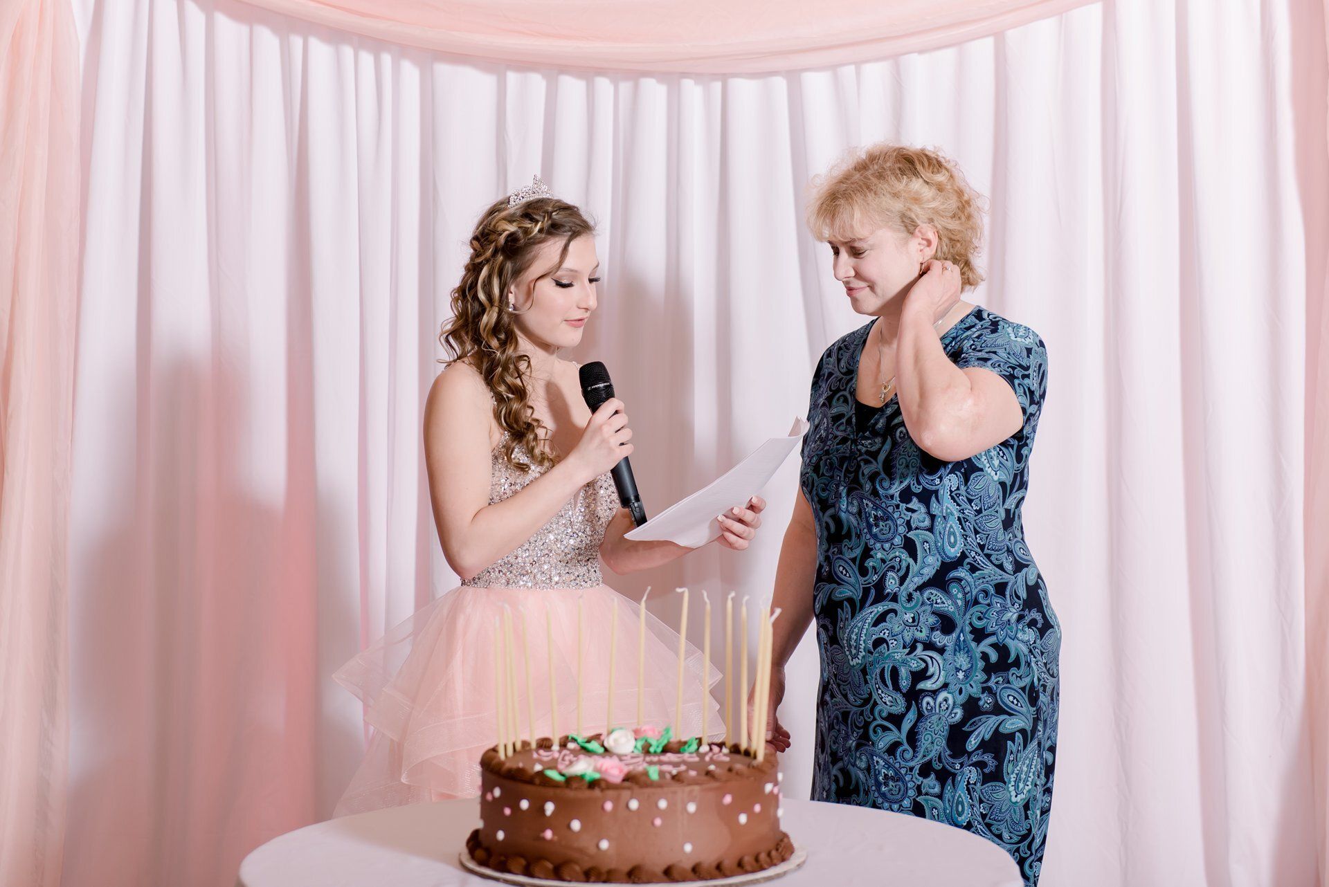 Two people at a birthday party beside a cake, one holding a microphone and the other touching their face.