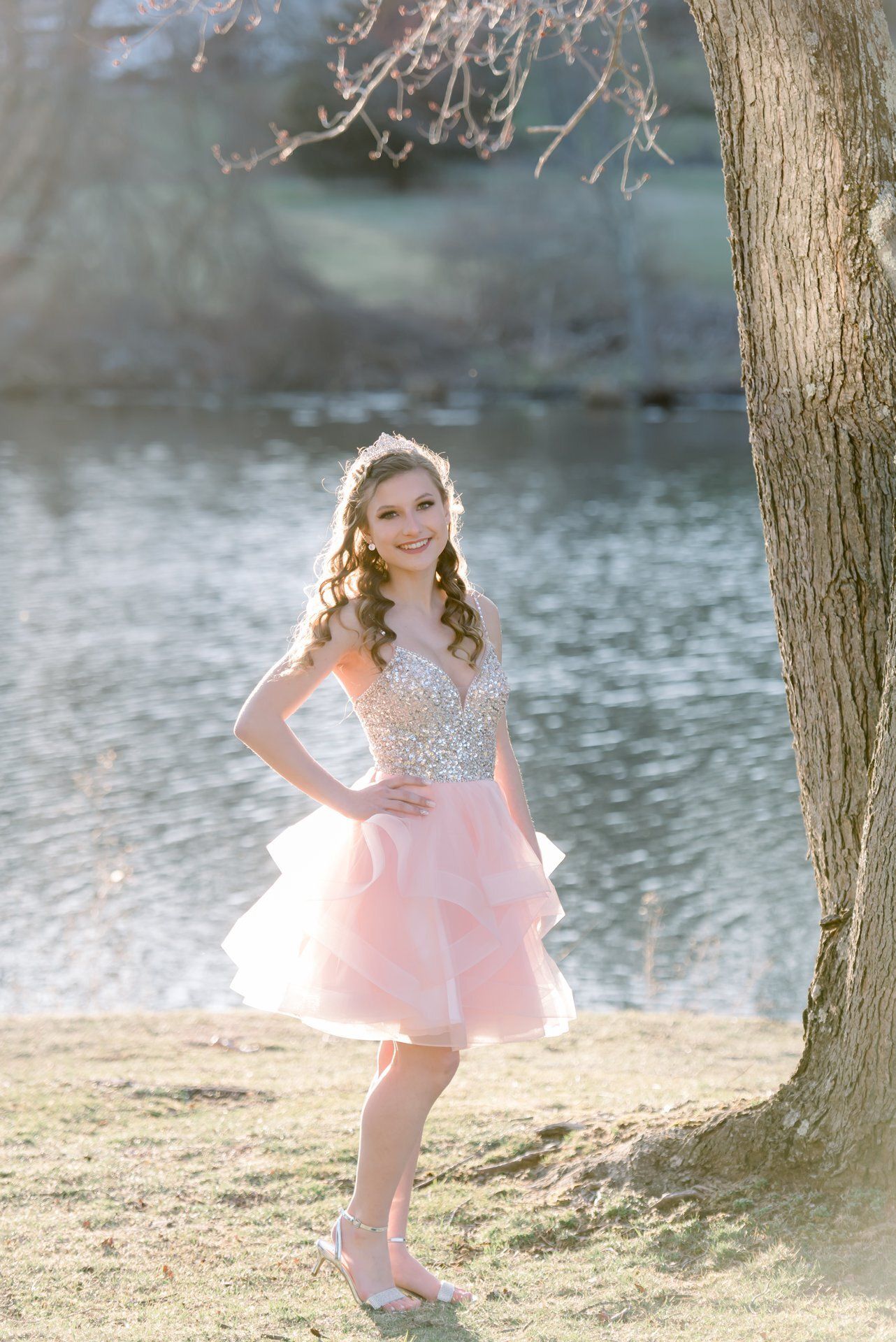 Person in a pink dress posing by a tree beside a lake on a sunny day
