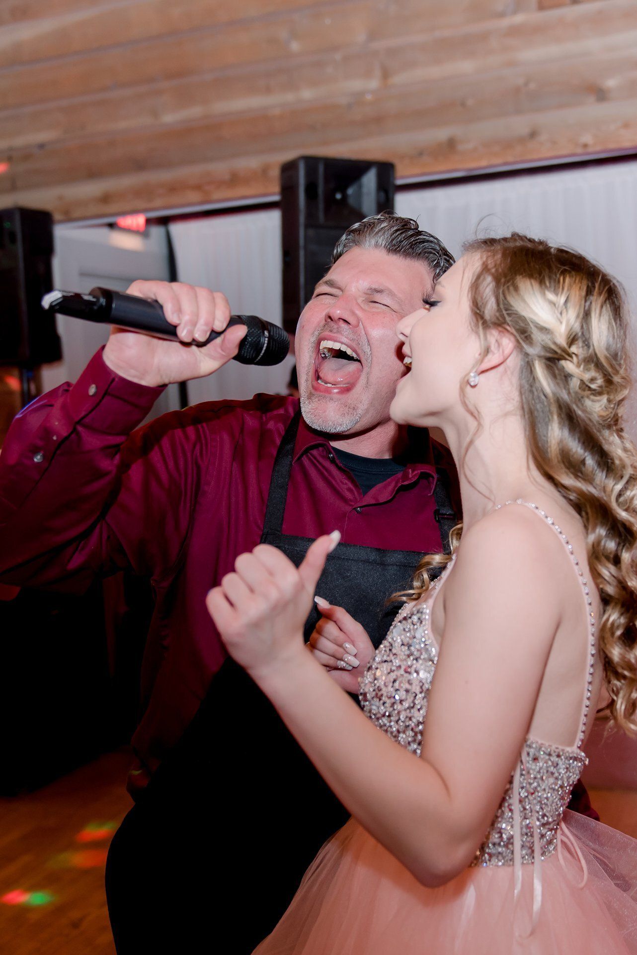 Man singing into a microphone while a woman in a pink dress stands beside him at an indoor event