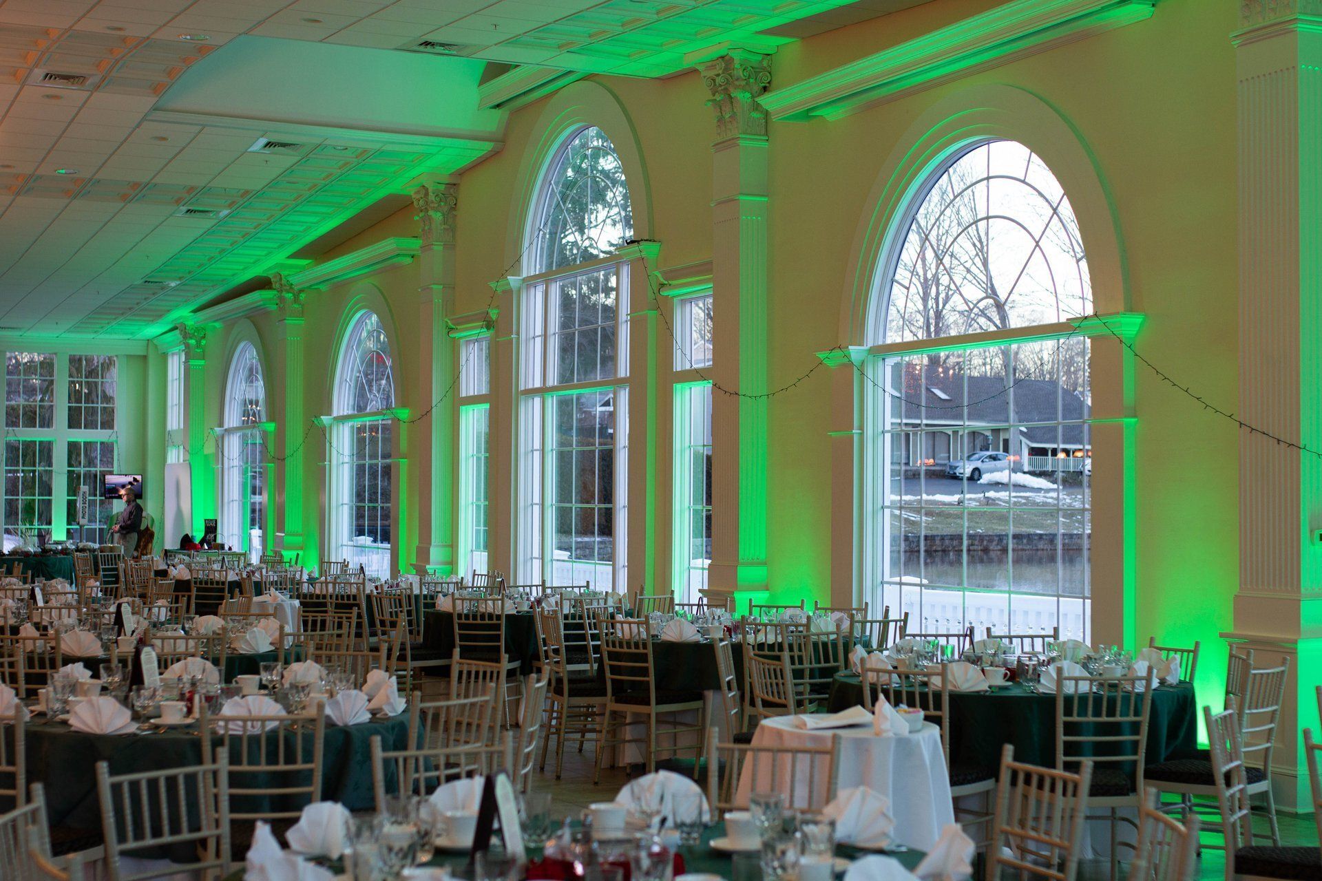 Banquet hall with round tables and chairs, lit by green uplights along tall arched windows.