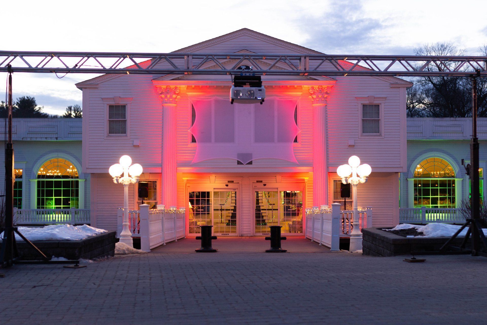 White building lit with pink and red lights at dusk, with a decorated entrance and lamp posts in front.