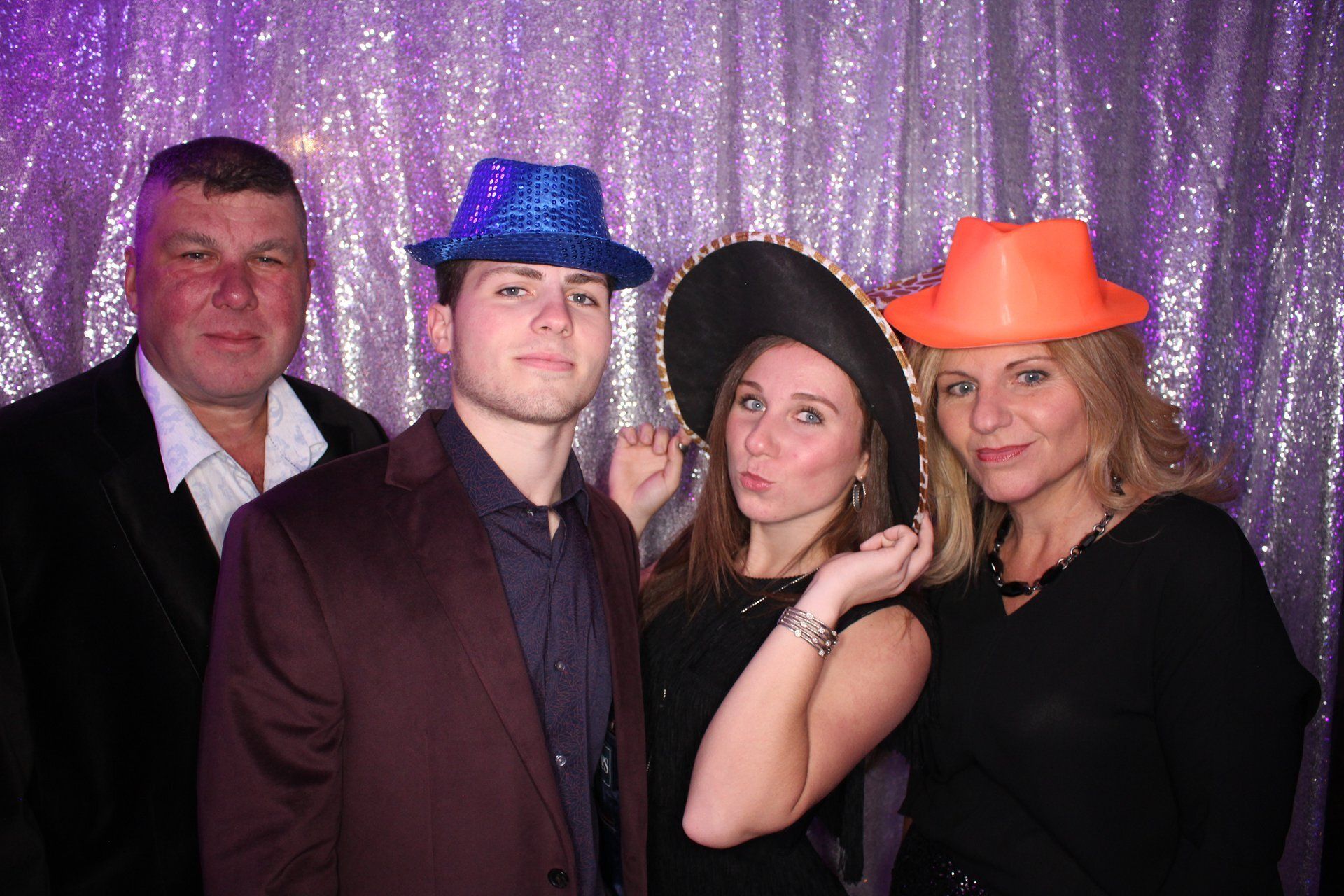 Four people posing in front of a purple sequin backdrop, wearing colorful party hats.