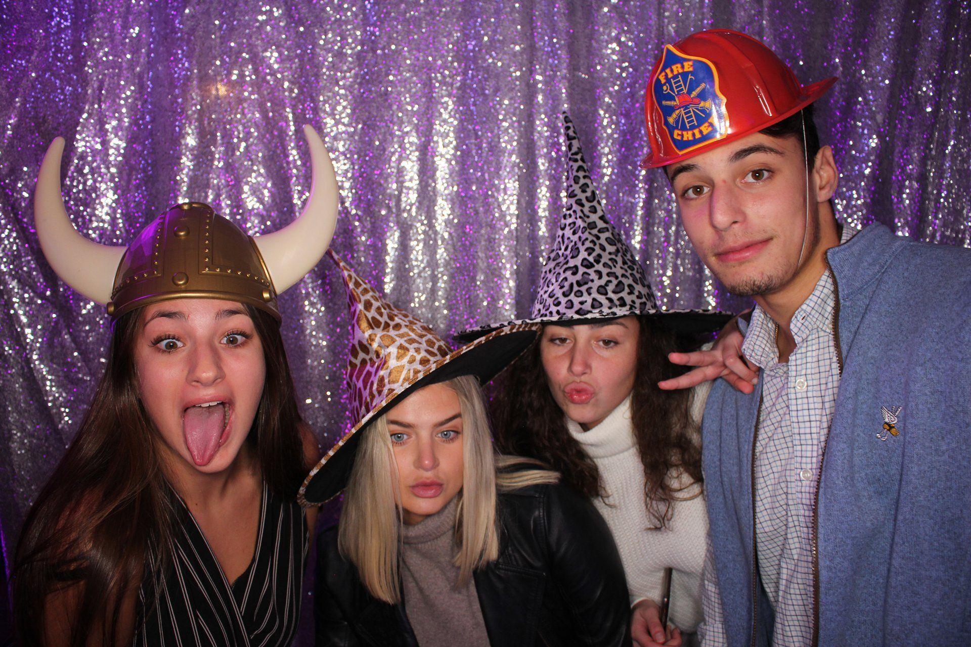 Four people wearing festive hats pose in front of a purple sequin backdrop, one making a playful face.