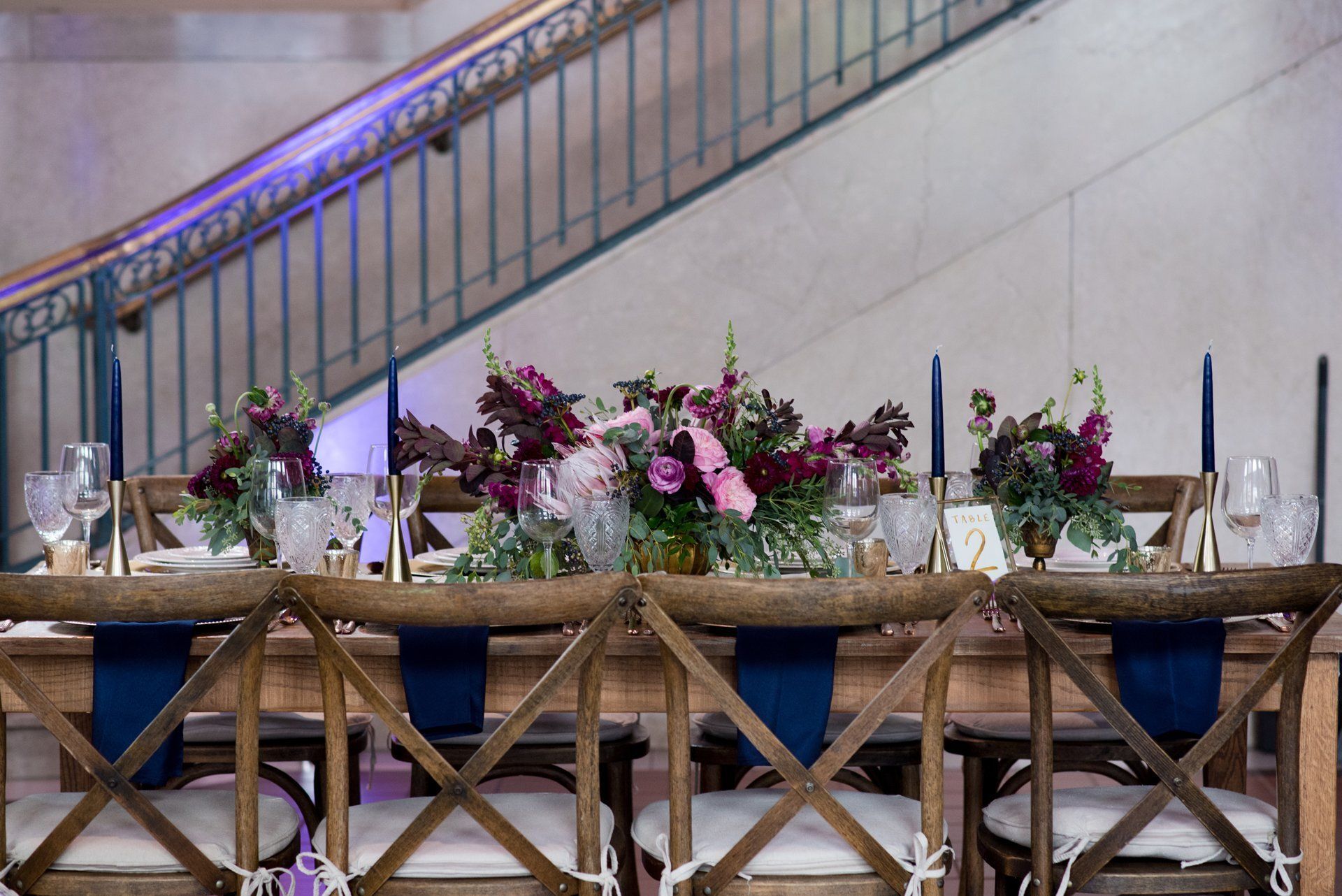 Elegant banquet table with white chairs, floral centerpieces, candles, and glassware beside a staircase.