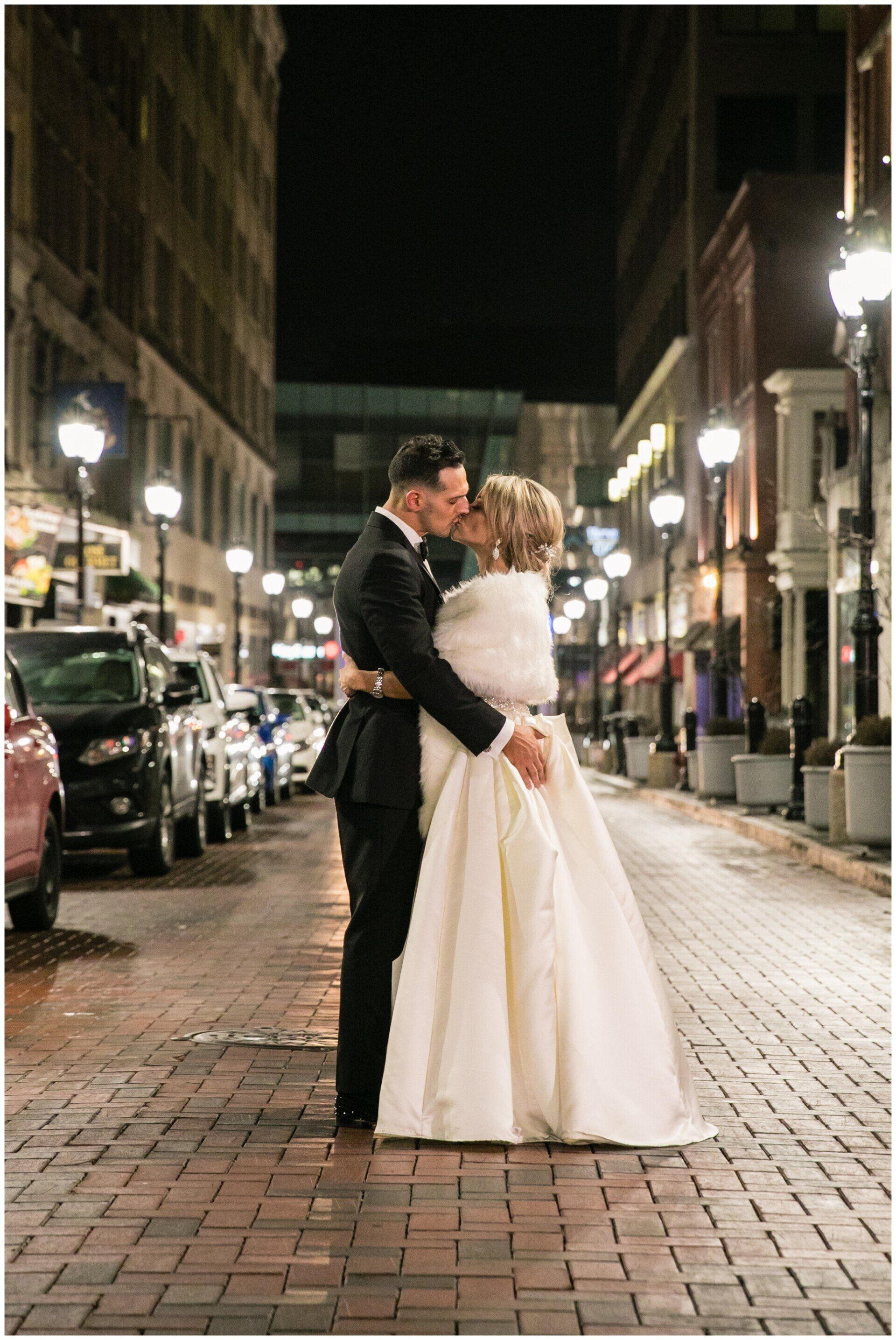 Couple embracing on a cobblestone street at night, city lights glowing around them.