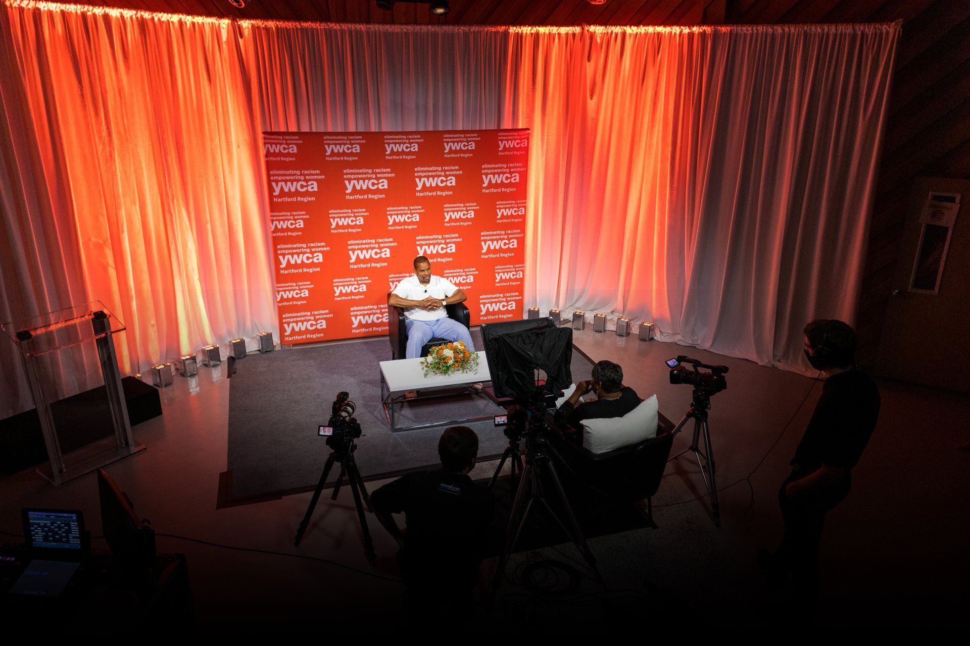 Press conference stage with red backdrop, white curtains, table, and several cameras in front