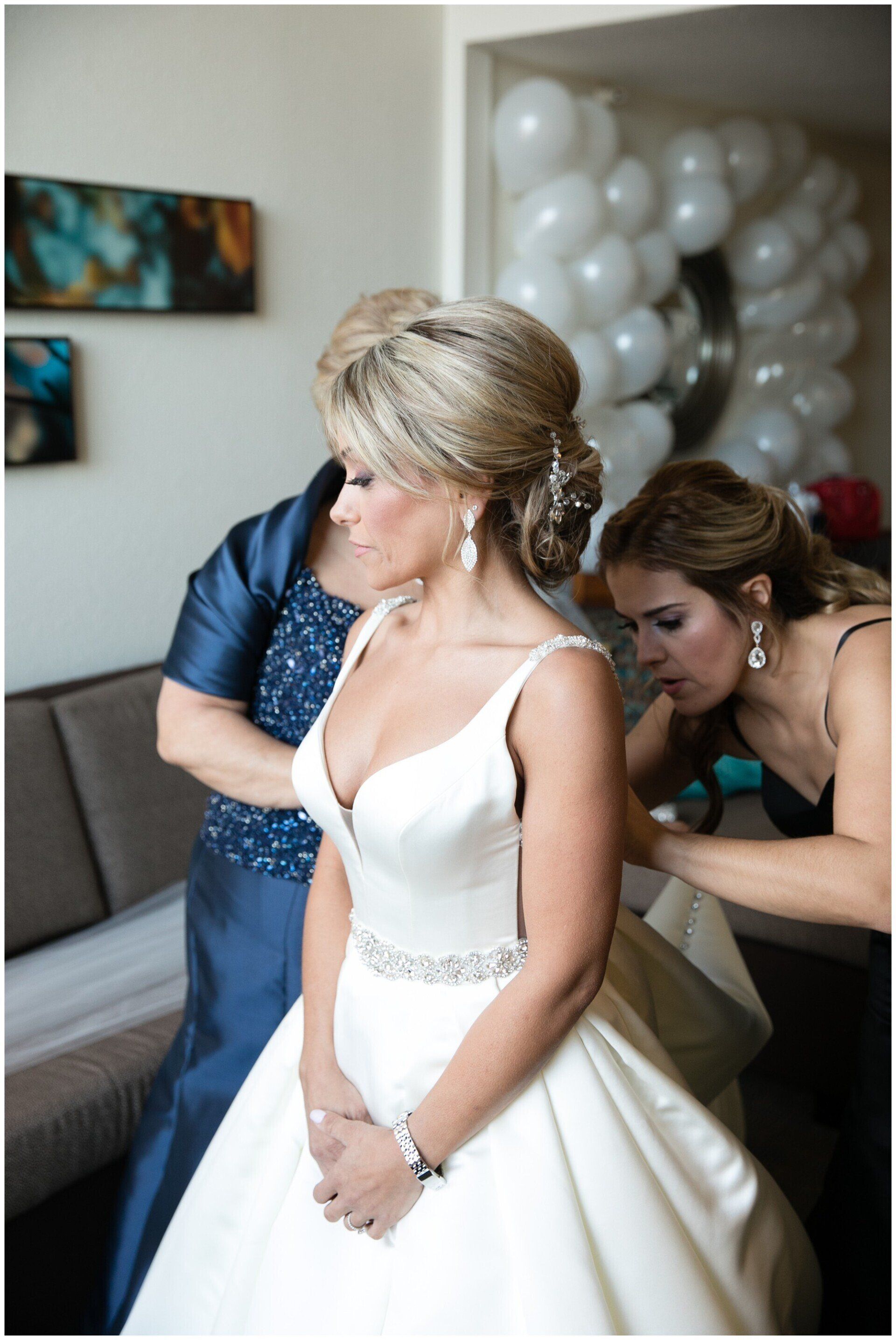 Bride in white gown being fastened by a helper in a room with white balloons