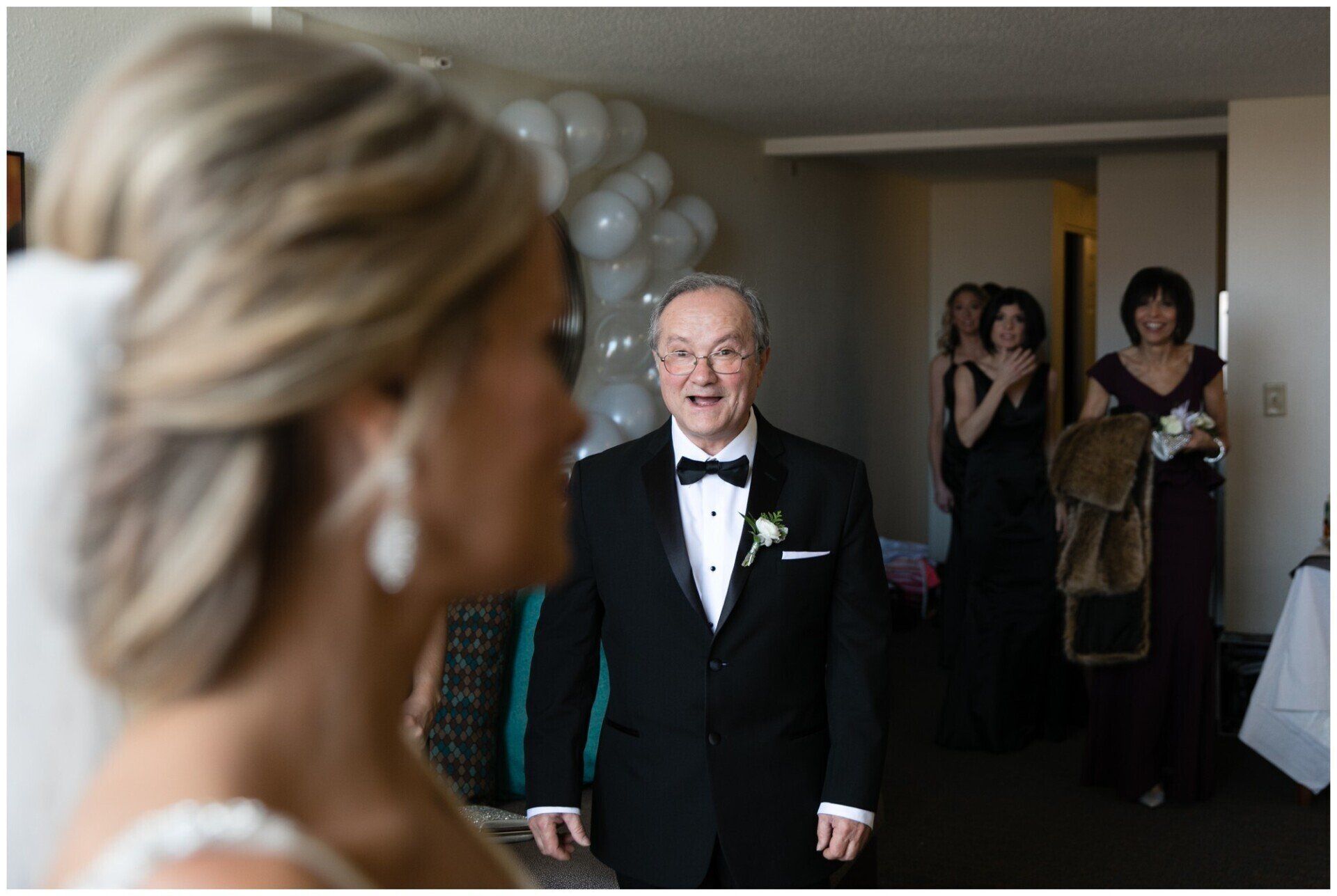 Bride in foreground watching an older man in a tuxedo smile at an indoor event entrance