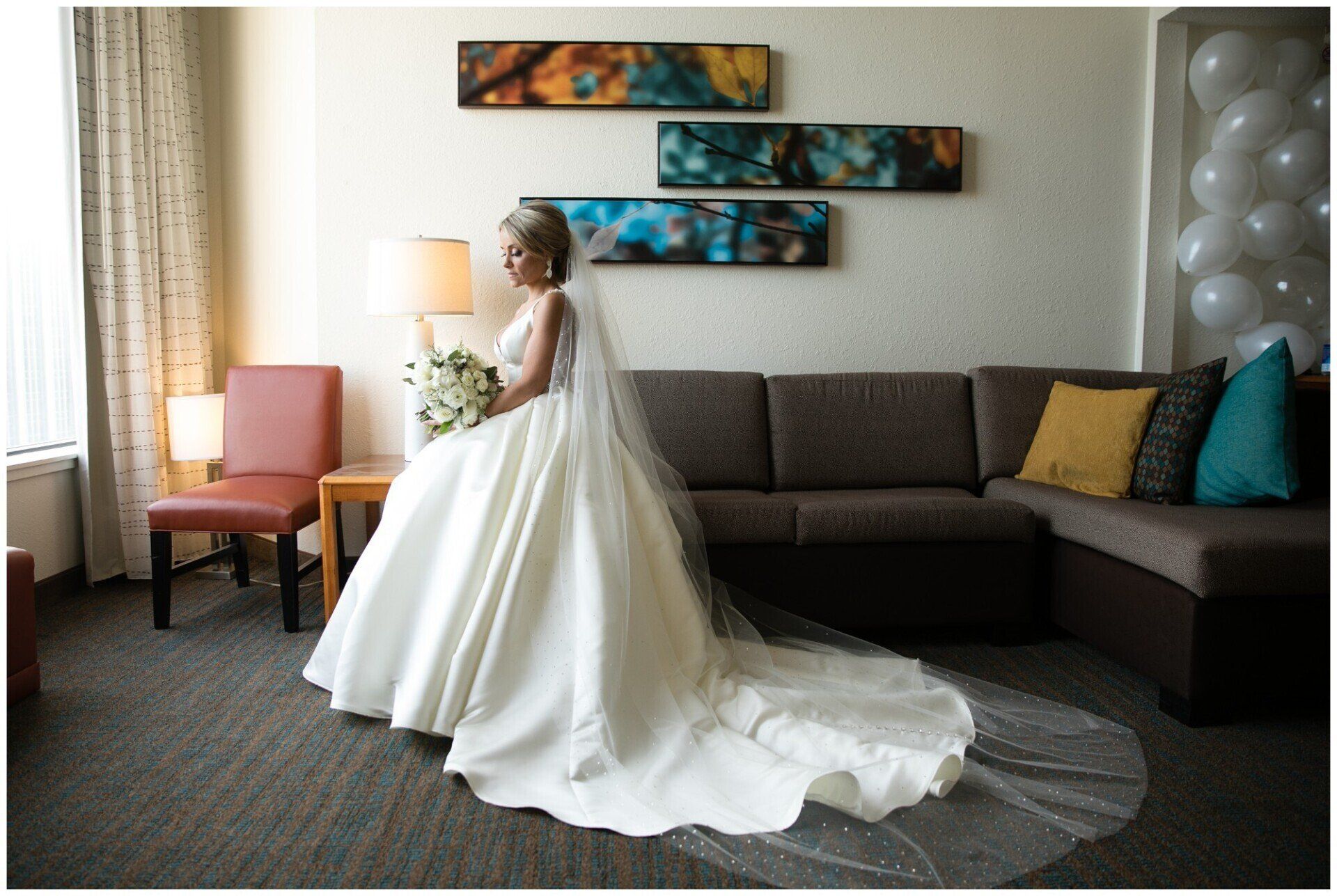 Bride in a white gown standing in a hotel room beside a dark sectional sofa and pink chair