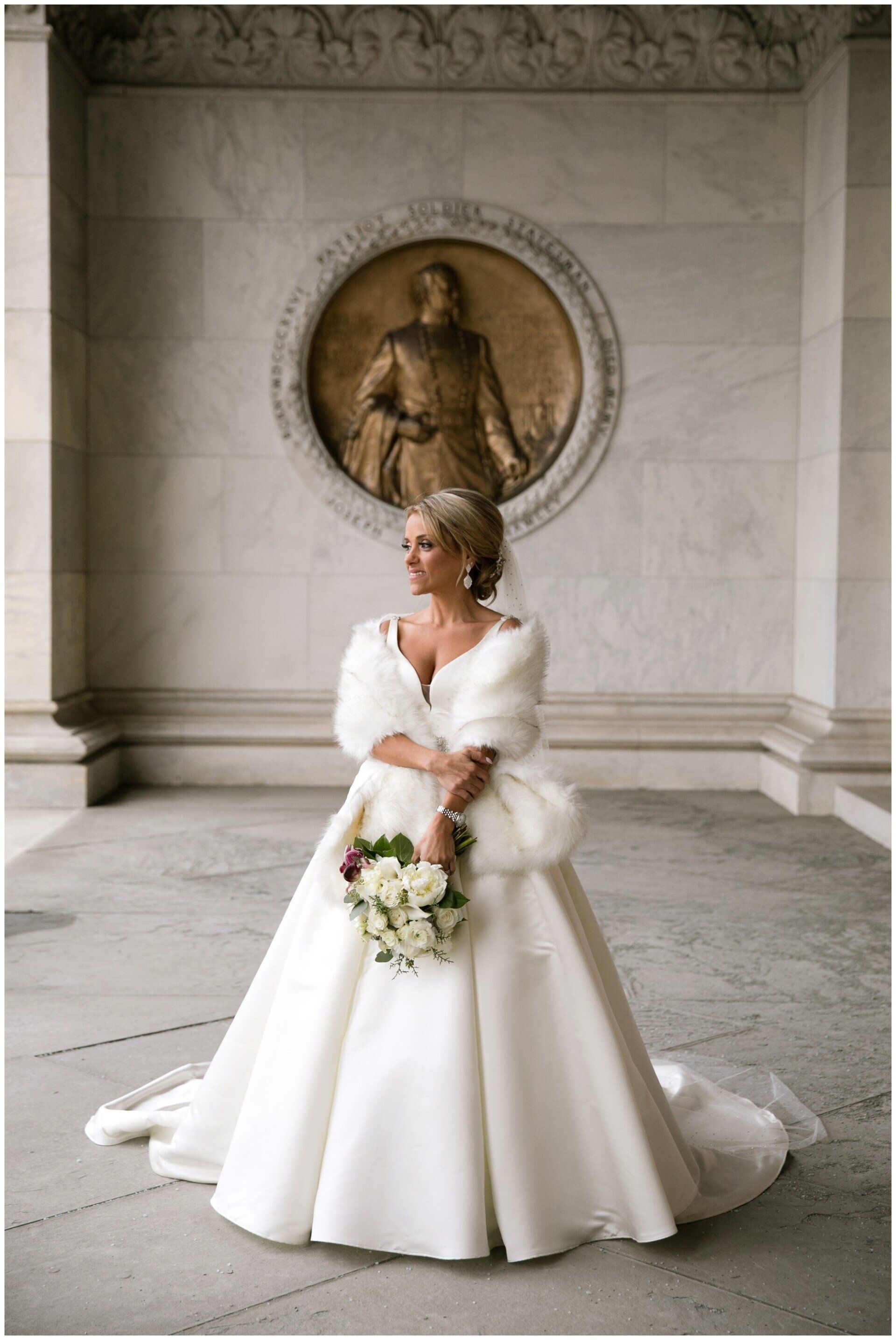 Bride in a white gown holding a bouquet in a marble hall before a round relief sculpture