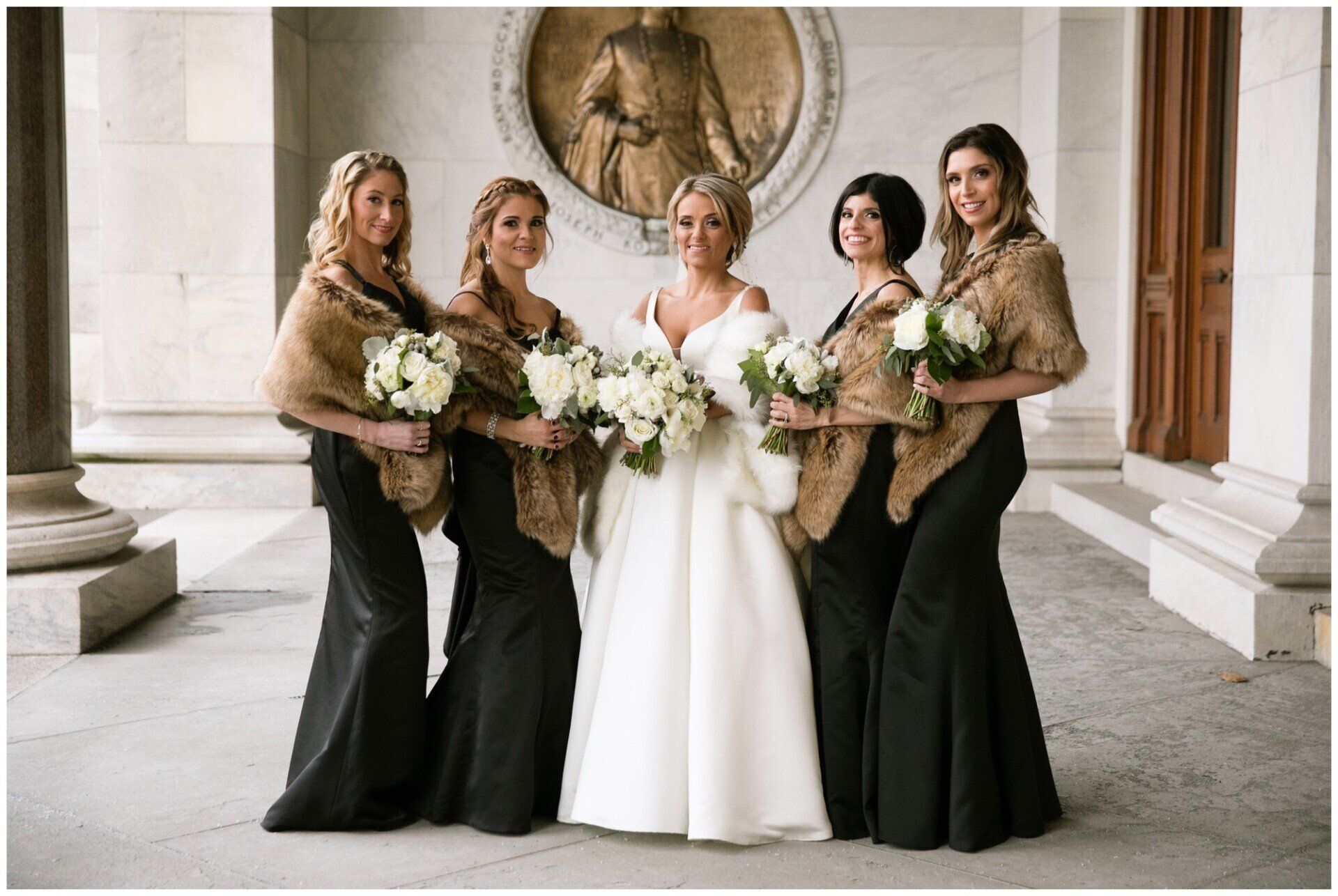 Five people in formal dresses hold white bouquets in a grand columned hall.