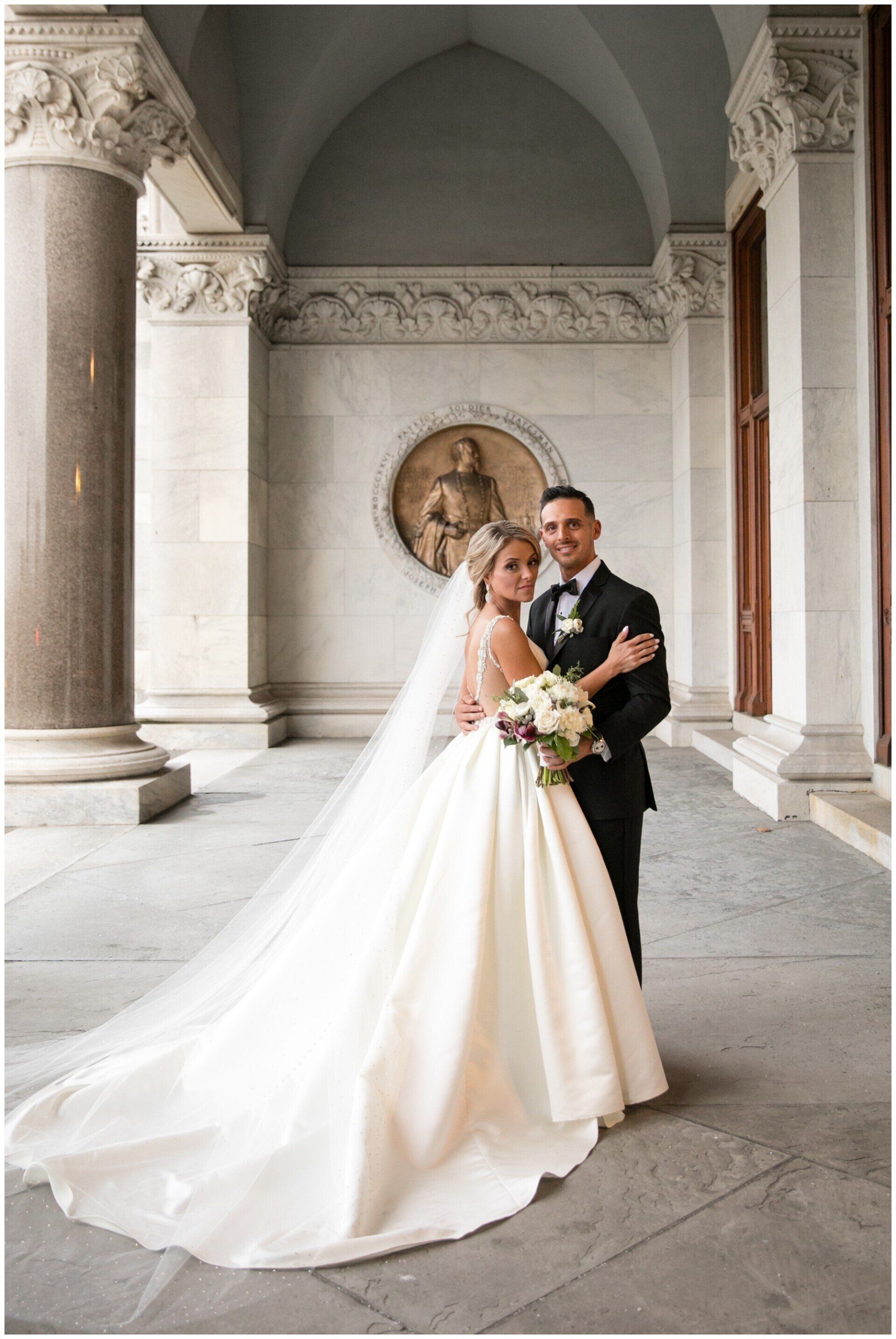 Bride and groom posing in a grand stone archway, bride in white gown with long veil and bouquet