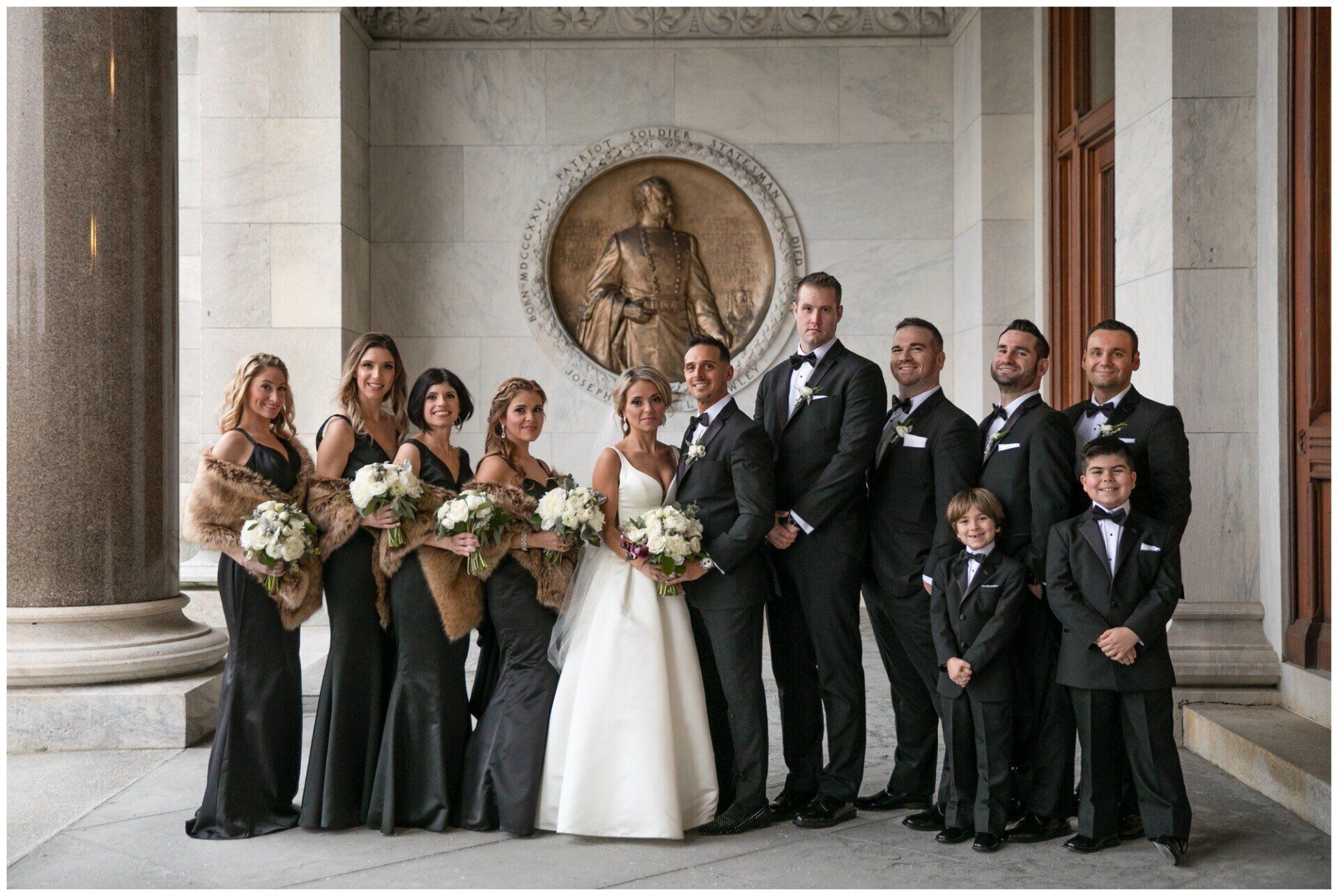 Wedding party posing in formal attire inside a stone building, with bride in white and bouquets.