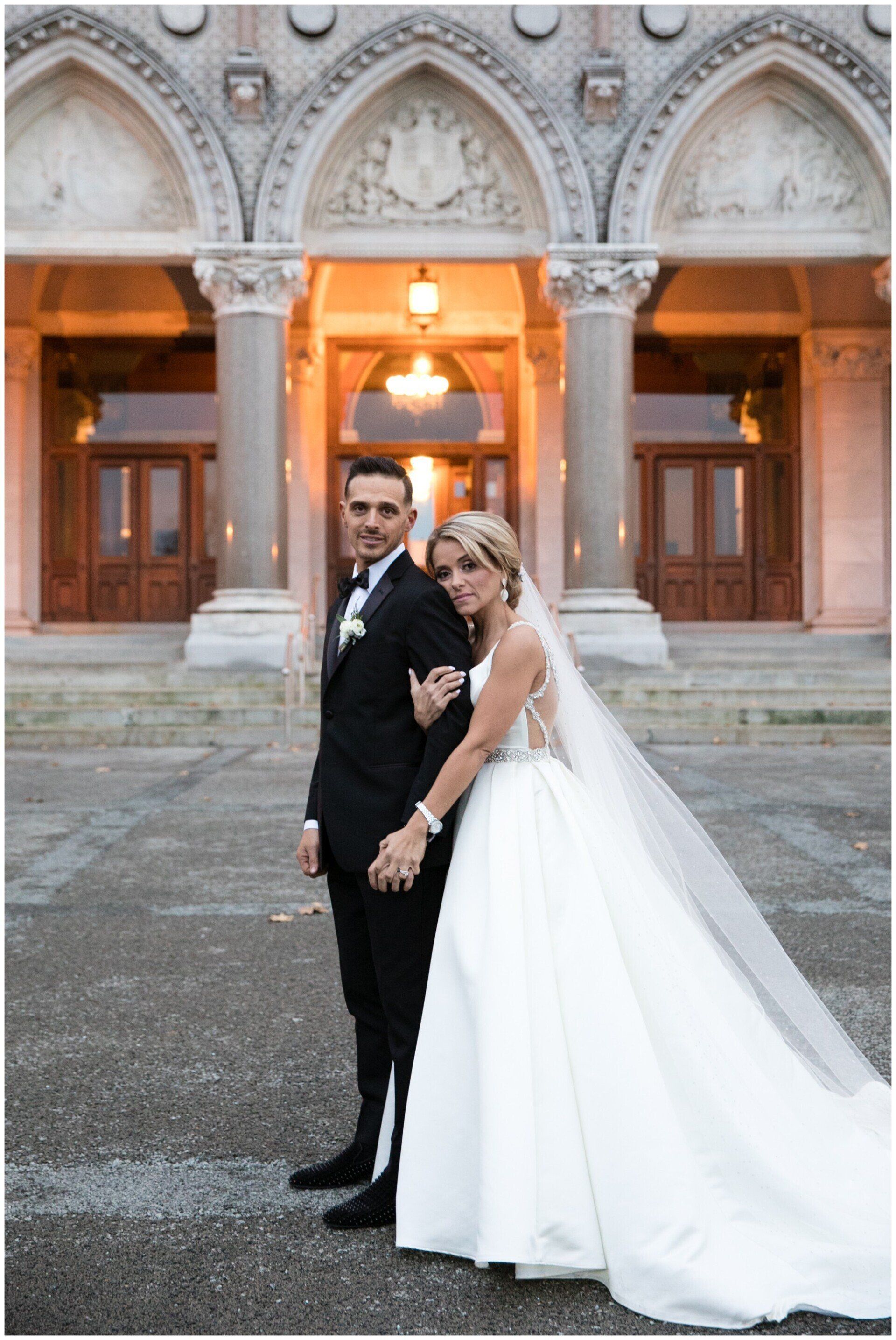 Bride and groom posing in wedding attire under ornate stone arches at a historic building