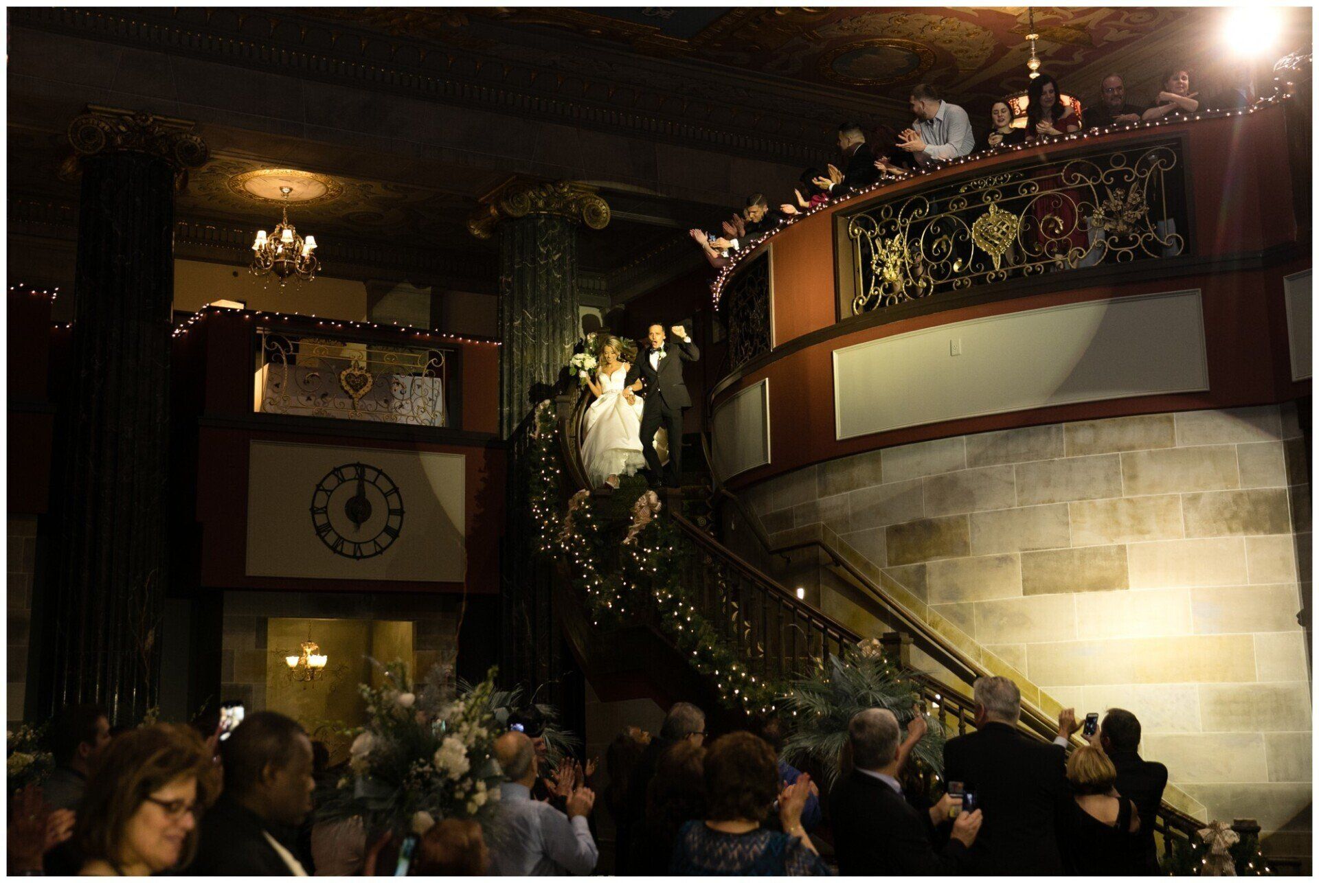 Crowded dimly lit grand hall with a staircase, ornate railings, and a lit statue at the landing