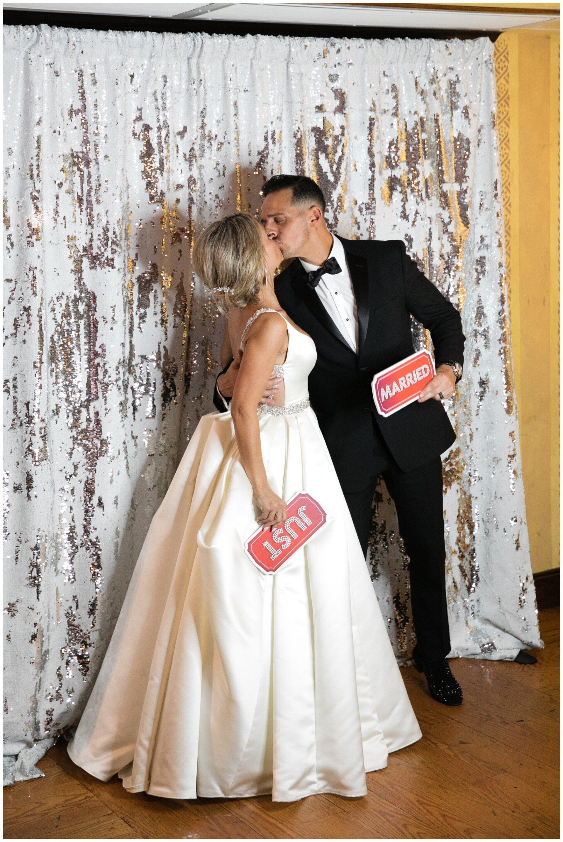 Bride and groom kissing in wedding attire, holding red props, against a silver sequin backdrop