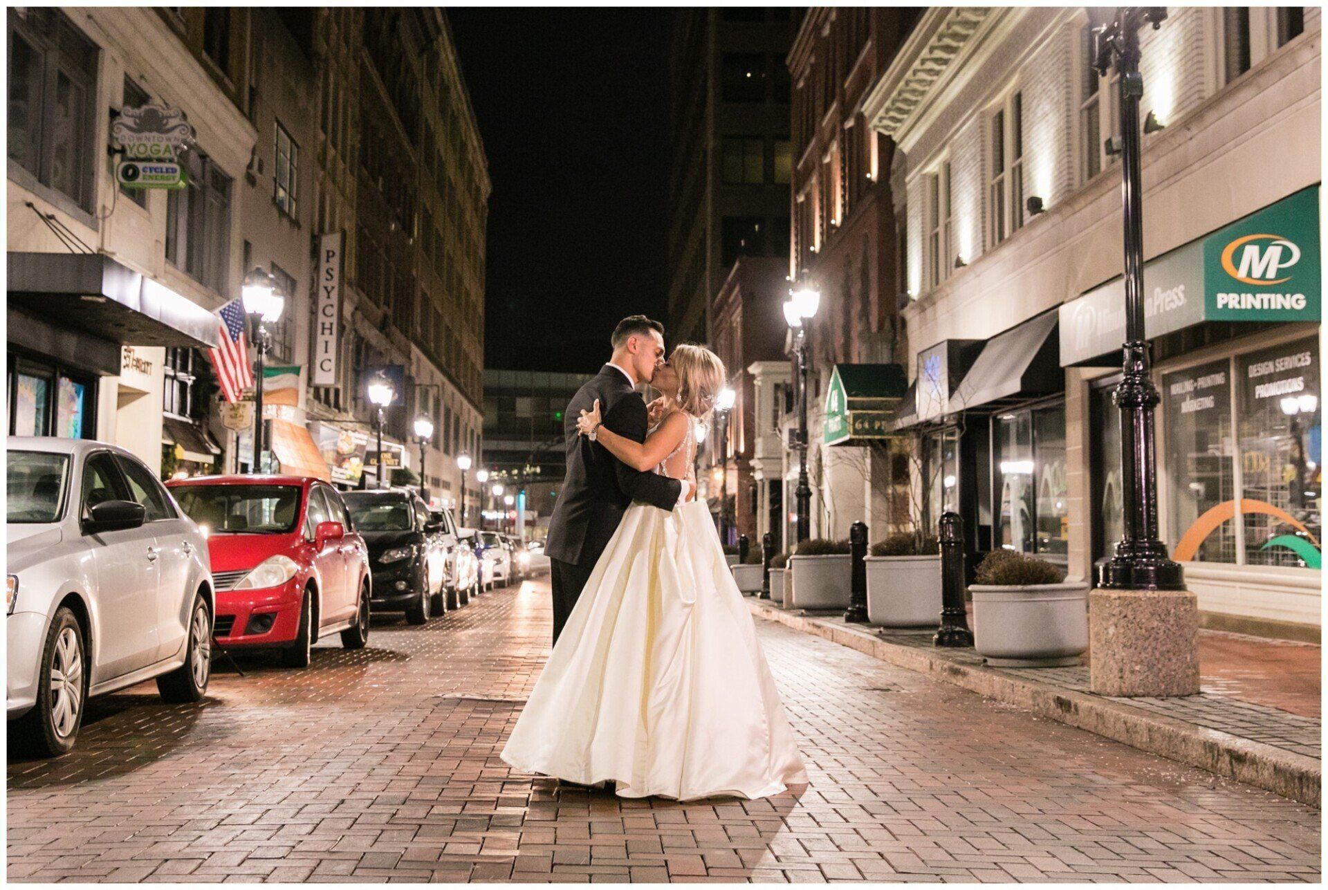 Couple in wedding attire embracing on a brick street at night, with parked cars and lit storefronts.