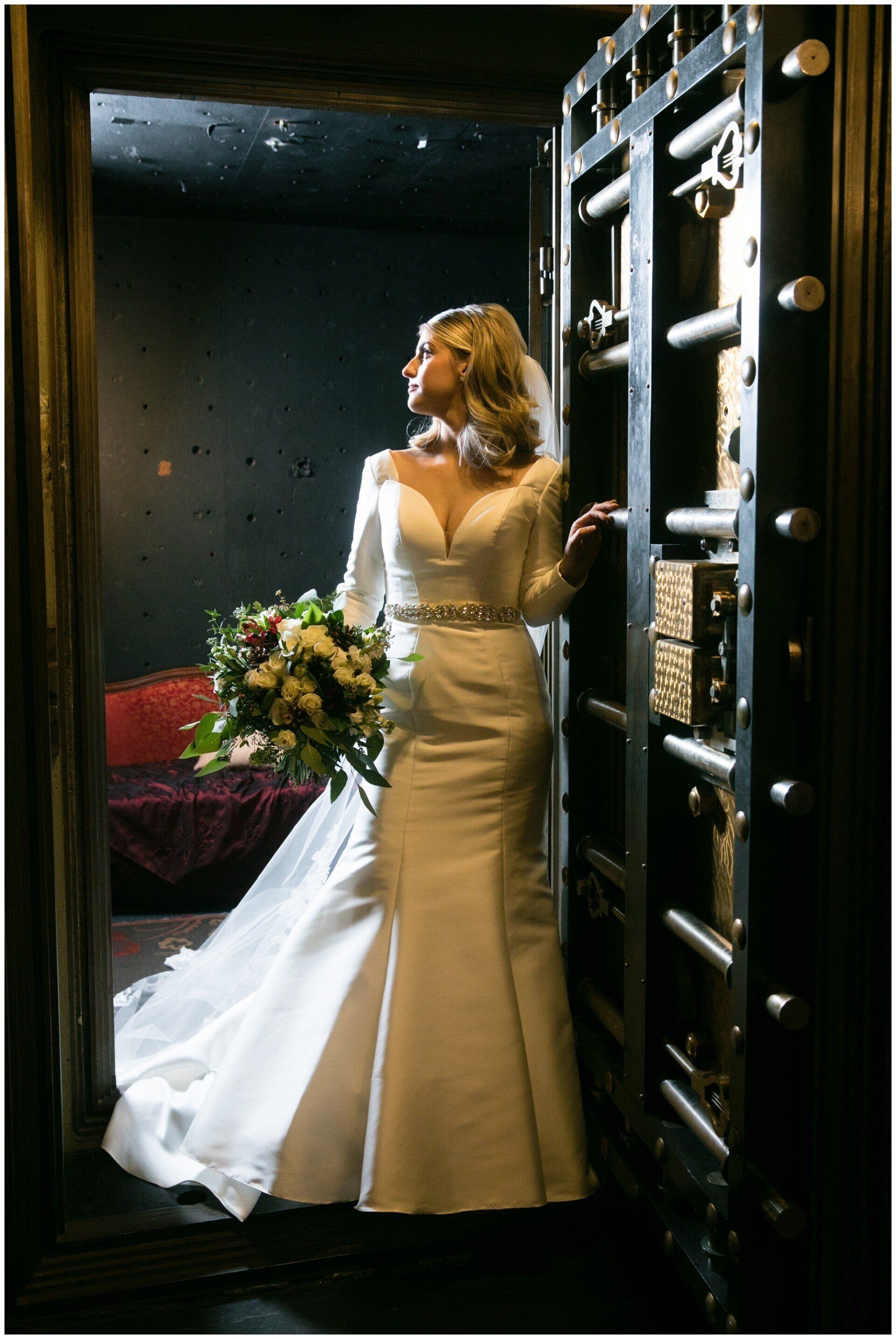 Woman in a white gown holding a bouquet, standing in a doorway beside a dark, ornate frame.