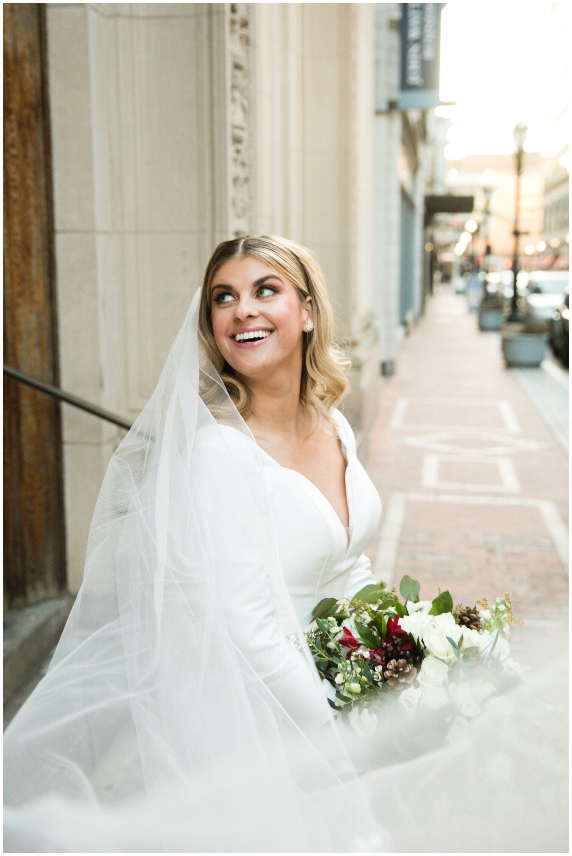 Bride in a white wedding dress holding a bouquet, smiling on a city sidewalk.