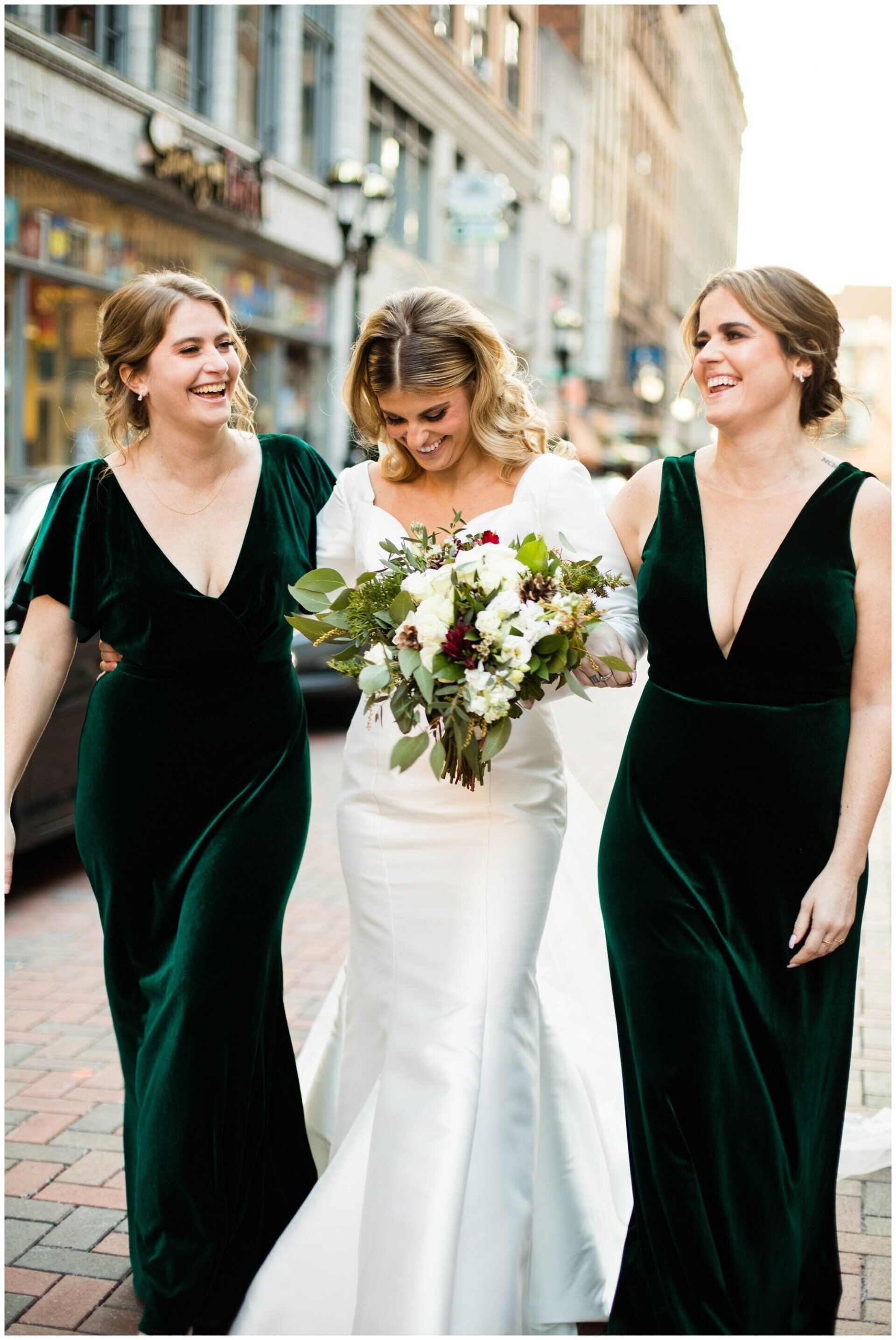 Bride in white dress walking with two women in dark green velvet dresses, smiling on a city street