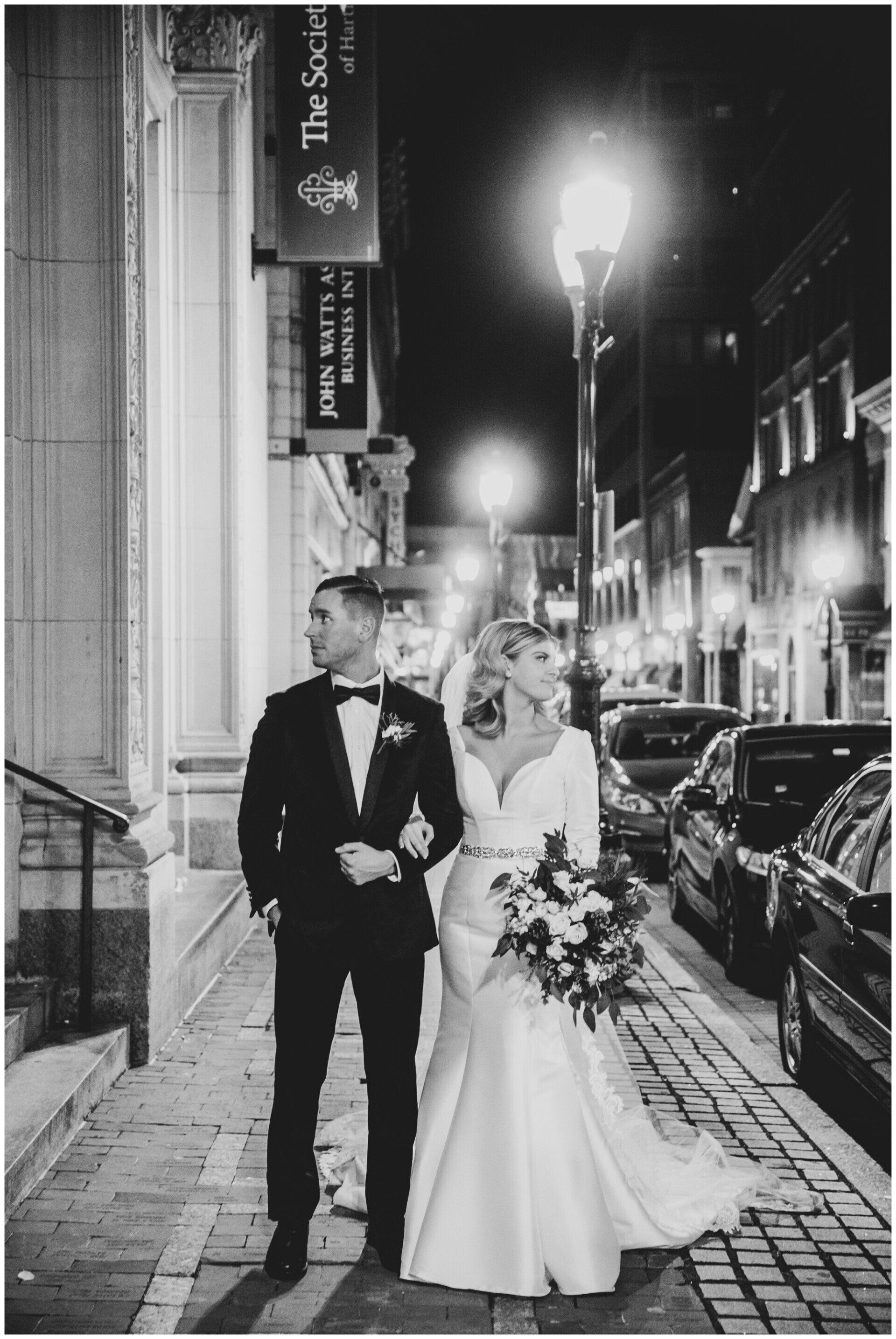 Newlyweds walking arm in arm on a city street at night, the bride holding a bouquet