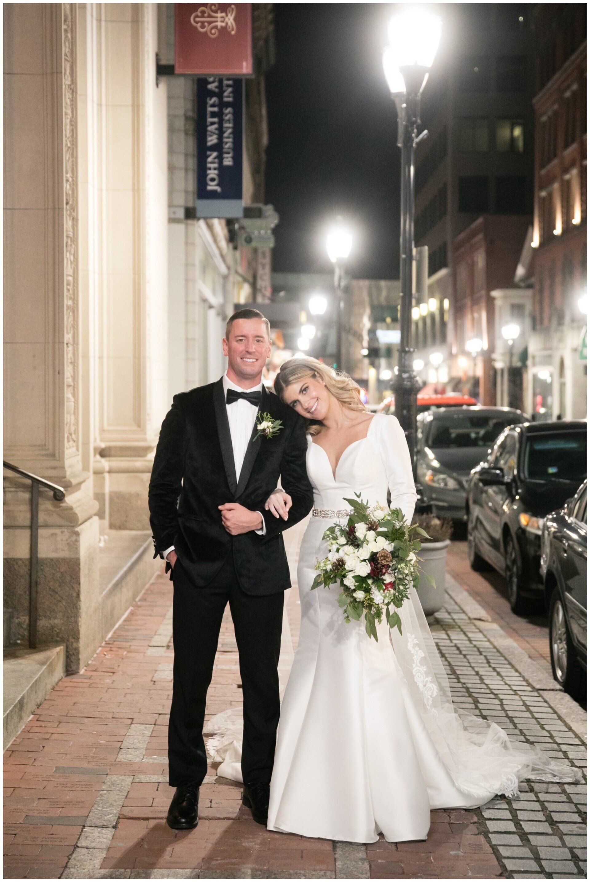 Bride and groom in formalwear smiling on a city sidewalk at night