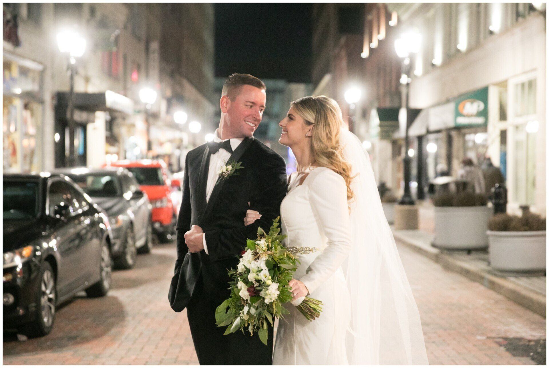 Bride and groom walking arm-in-arm on a warmly lit city street at night, smiling at each other