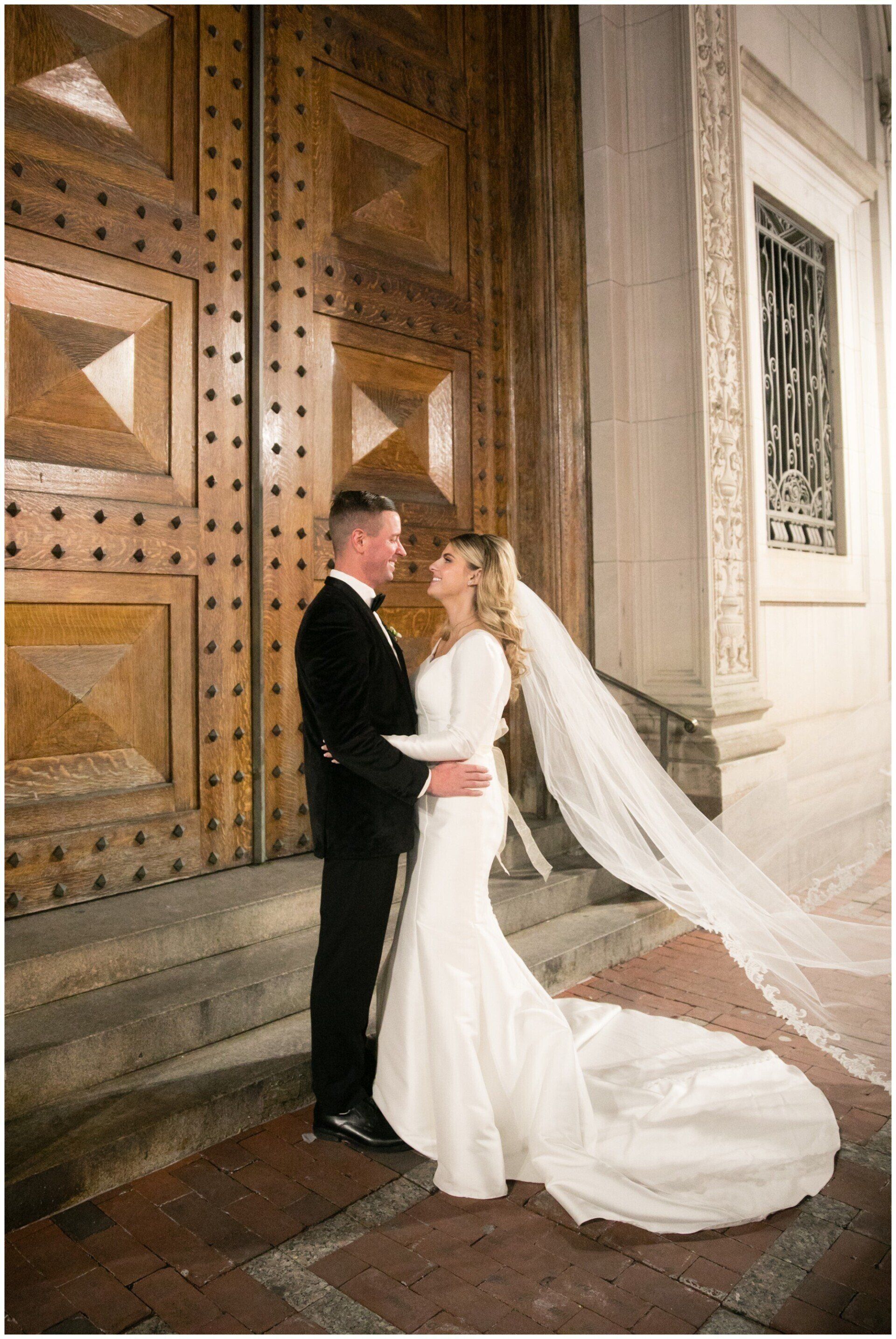 Bride and groom in wedding attire standing by ornate wooden doors, her veil trailing on the stone floor