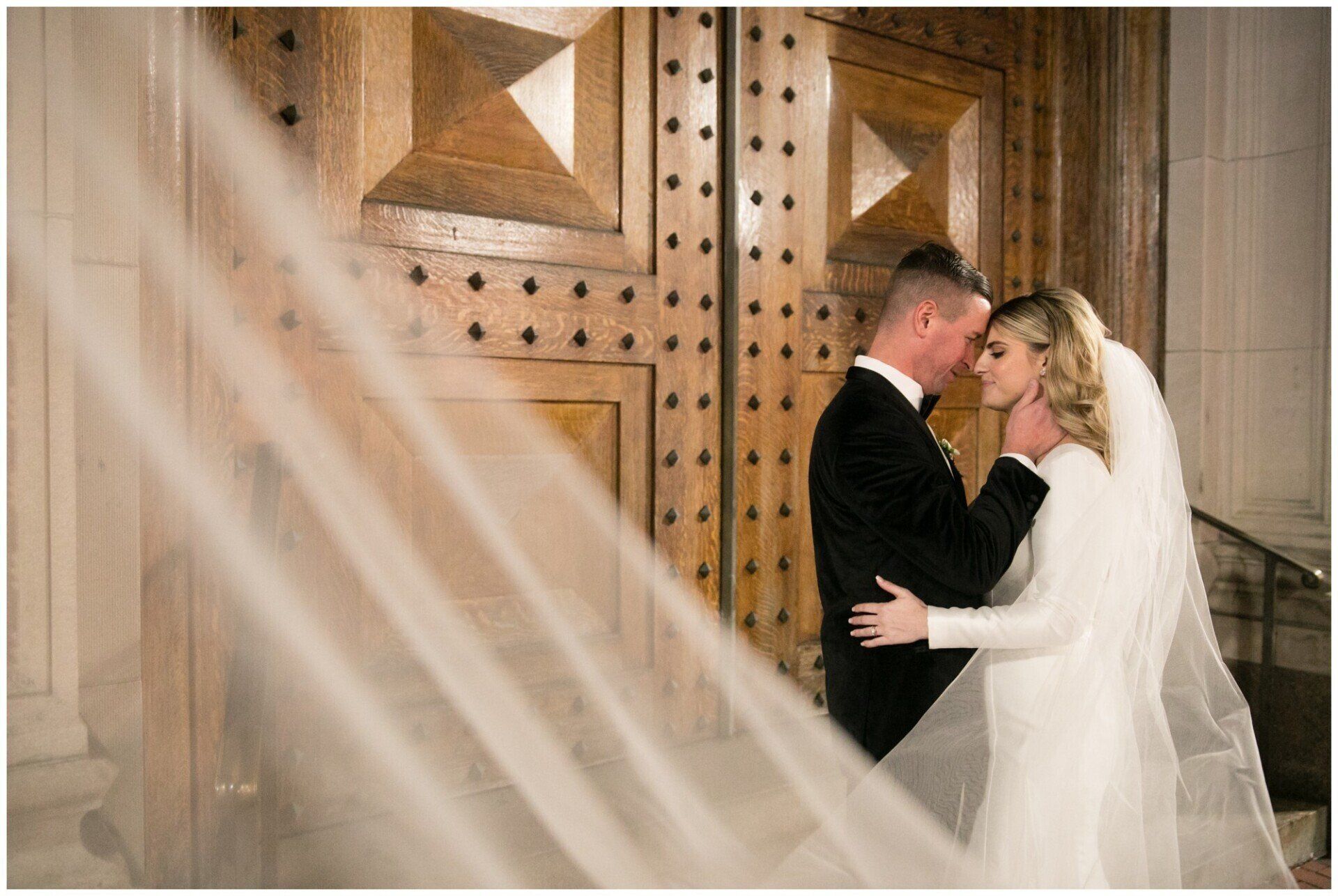 Bride and groom embracing before ornate wooden doors, with soft white fabric in the foreground