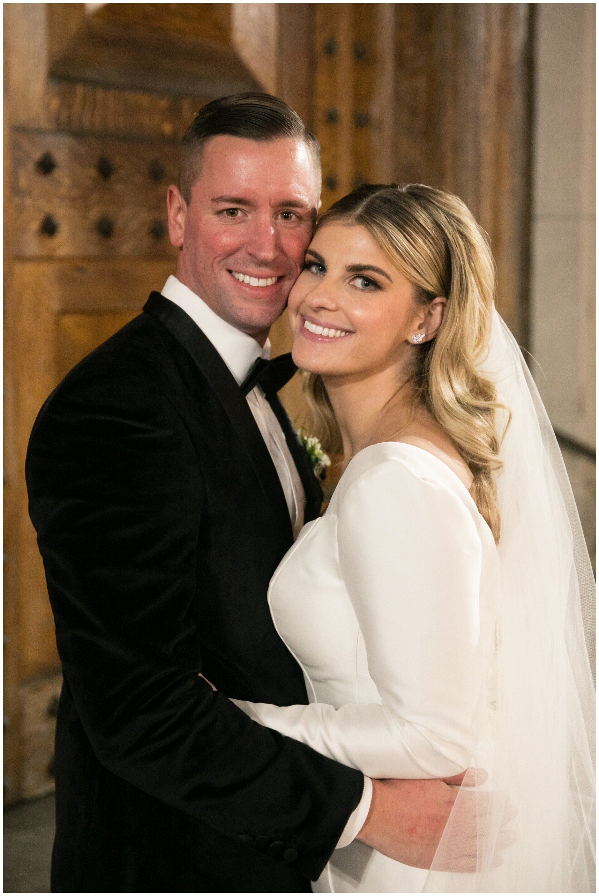 Smiling couple in formal wedding attire posing indoors, embracing in front of a wooden wall.