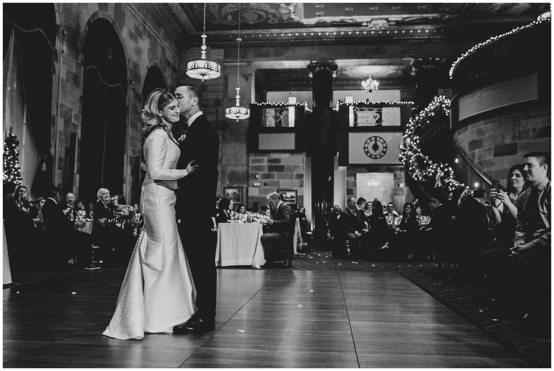 Black-and-white photo of a couple slow dancing in a decorated hall at a formal event.