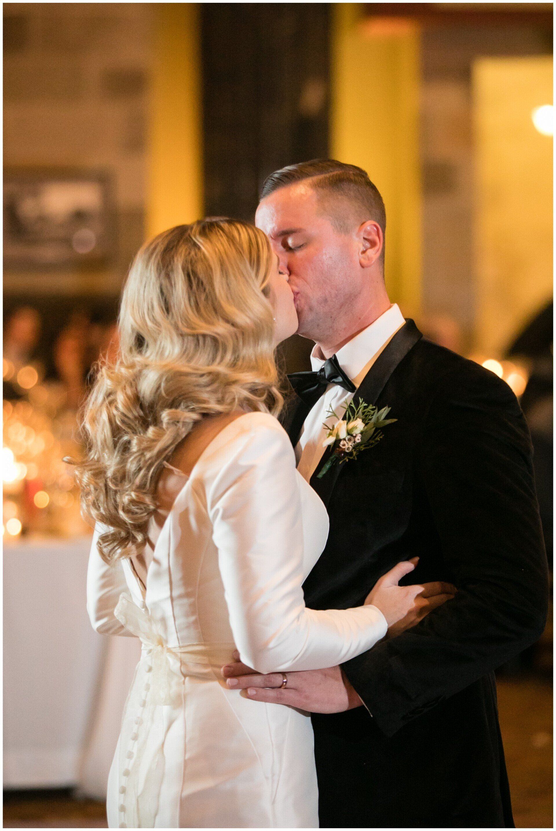 Couple in formal attire kissing at a warmly lit wedding reception