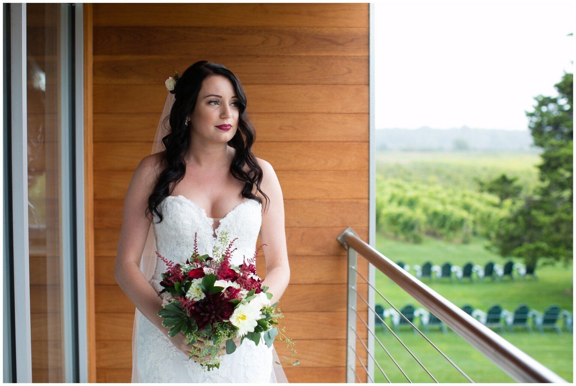 Bride in a white dress holding a red and white bouquet on a porch with a scenic green field behind her