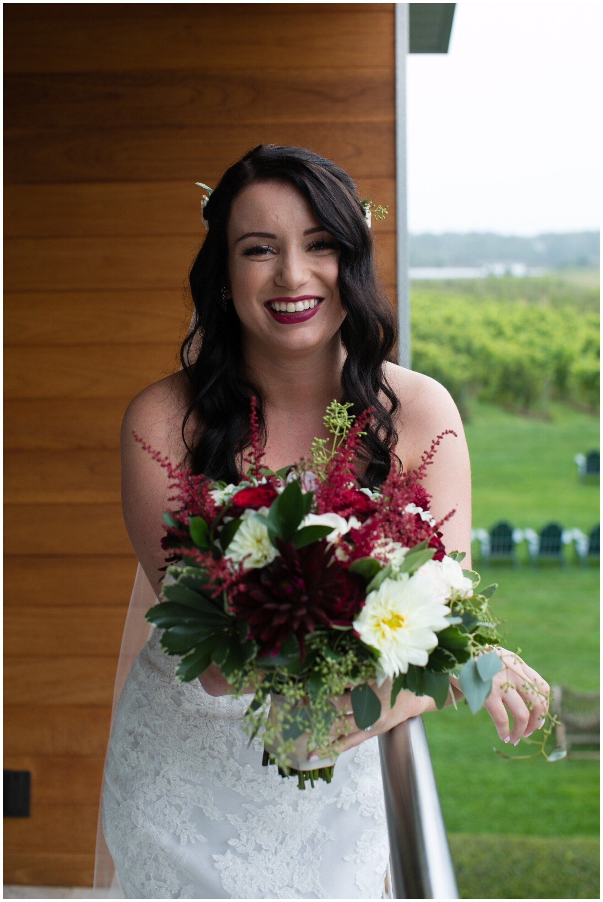 Bride in a white dress smiling on a balcony, holding a red and white bouquet.
