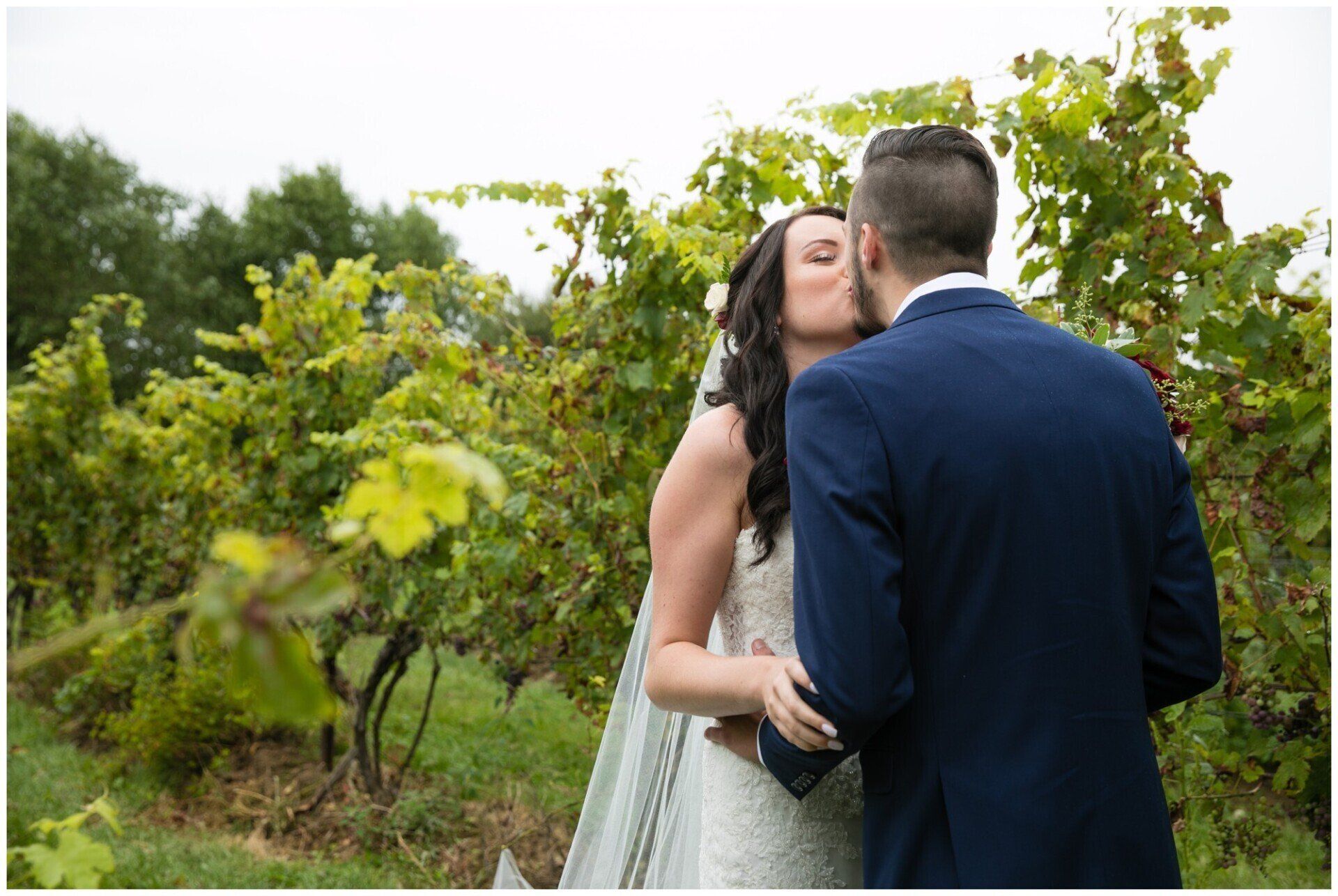 Couple kissing in a green vineyard, with the man in a navy suit and the woman in a white dress.