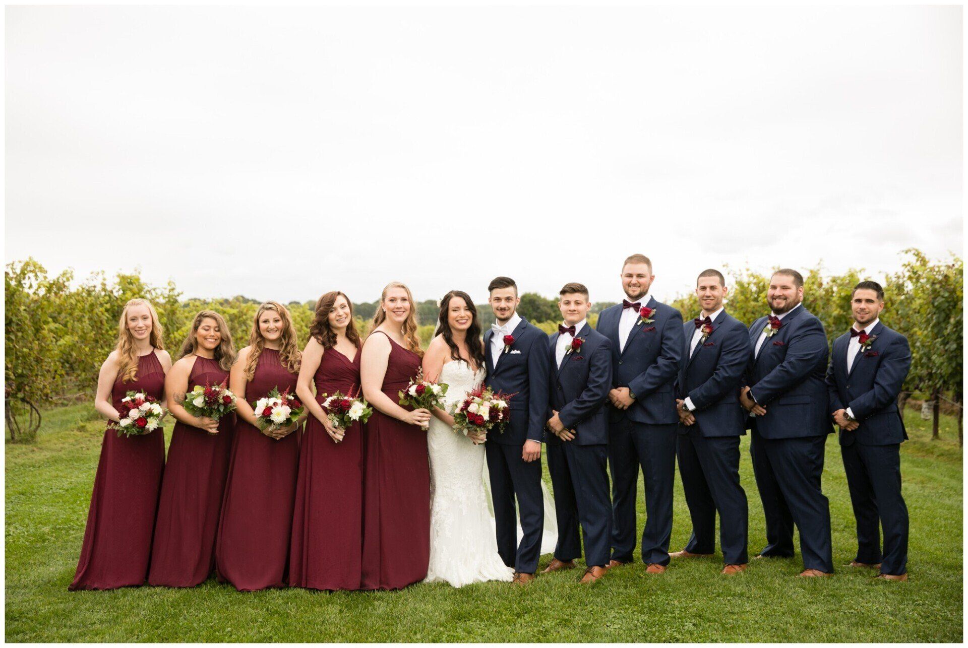 Wedding party posing outdoors on grass, with bridesmaids in burgundy dresses and groomsmen in navy suits