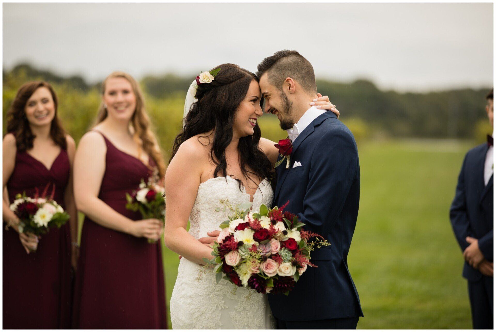 Bride and groom kiss outdoors, with bridesmaids in burgundy dresses behind them.