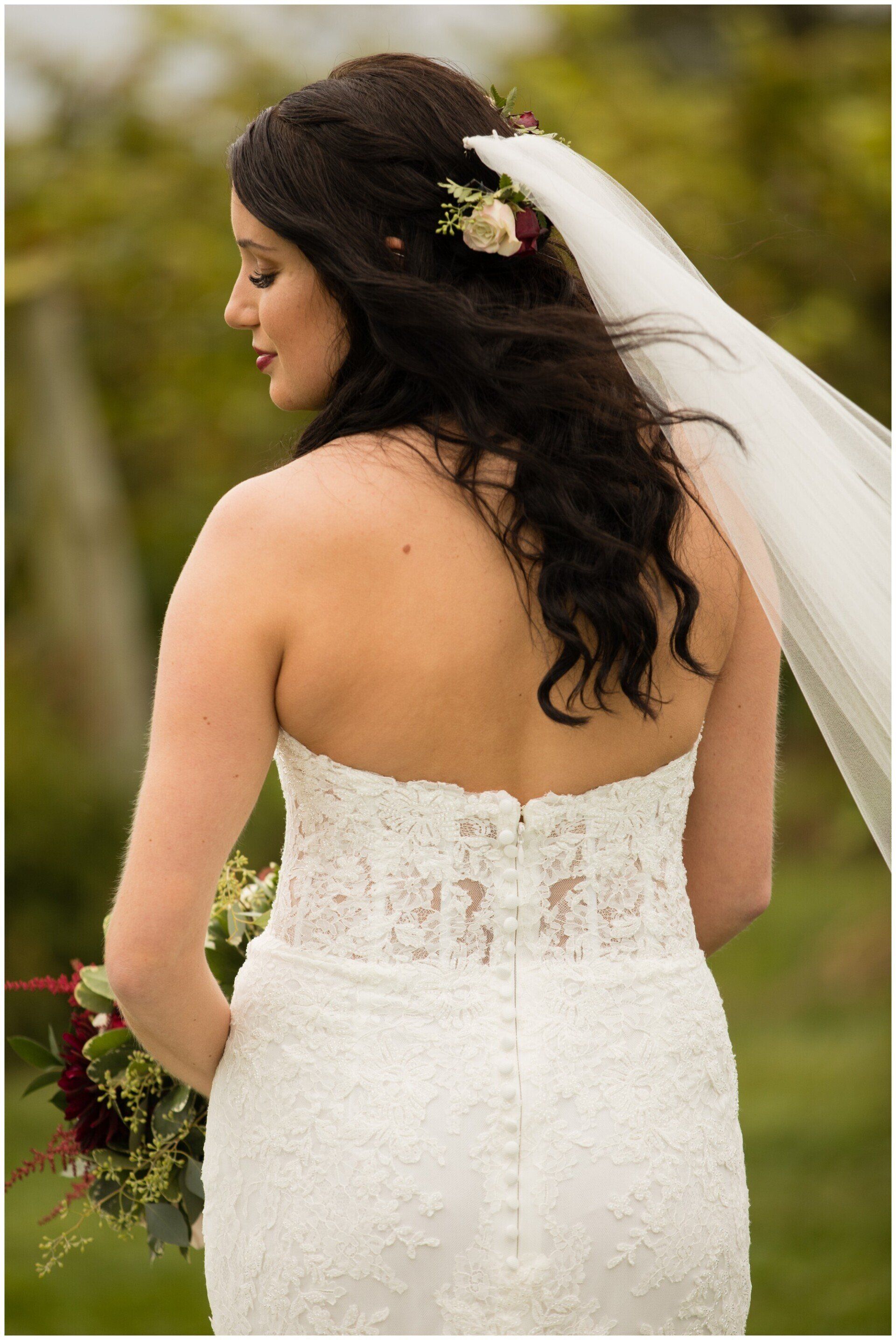 Bride in a white lace wedding dress and veil, seen from behind outdoors with flowers in her hair