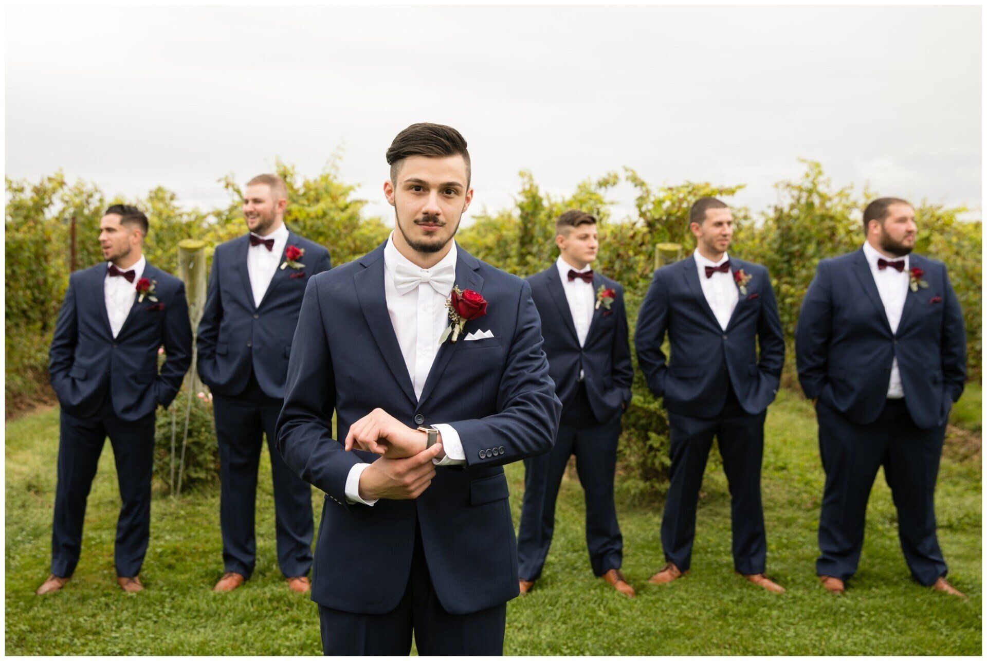 Six men in dark suits and red bow ties standing on grass outdoors, with one in front looking at the camera