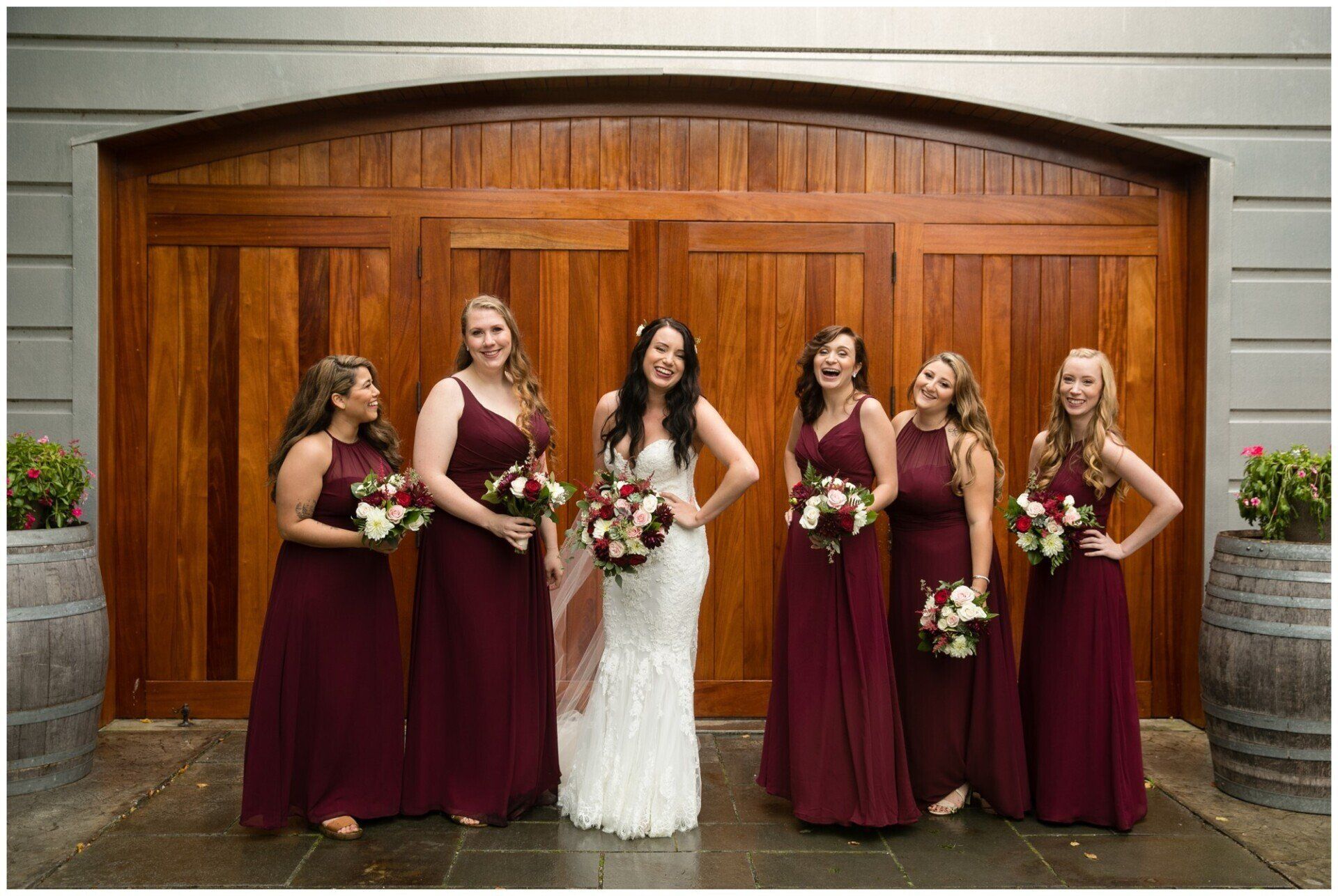 Five bridesmaids and a bride holding bouquets stand before wooden barn doors.