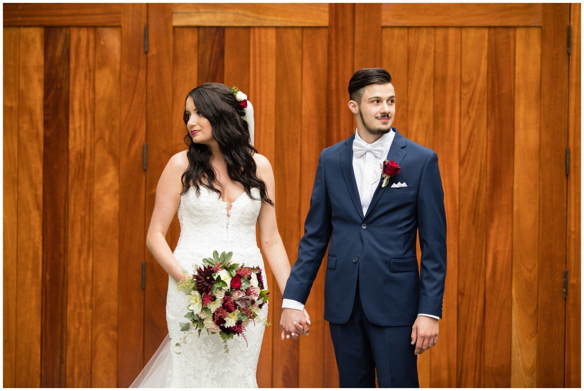 Bride and groom holding hands in front of a wooden backdrop, both dressed for a wedding.