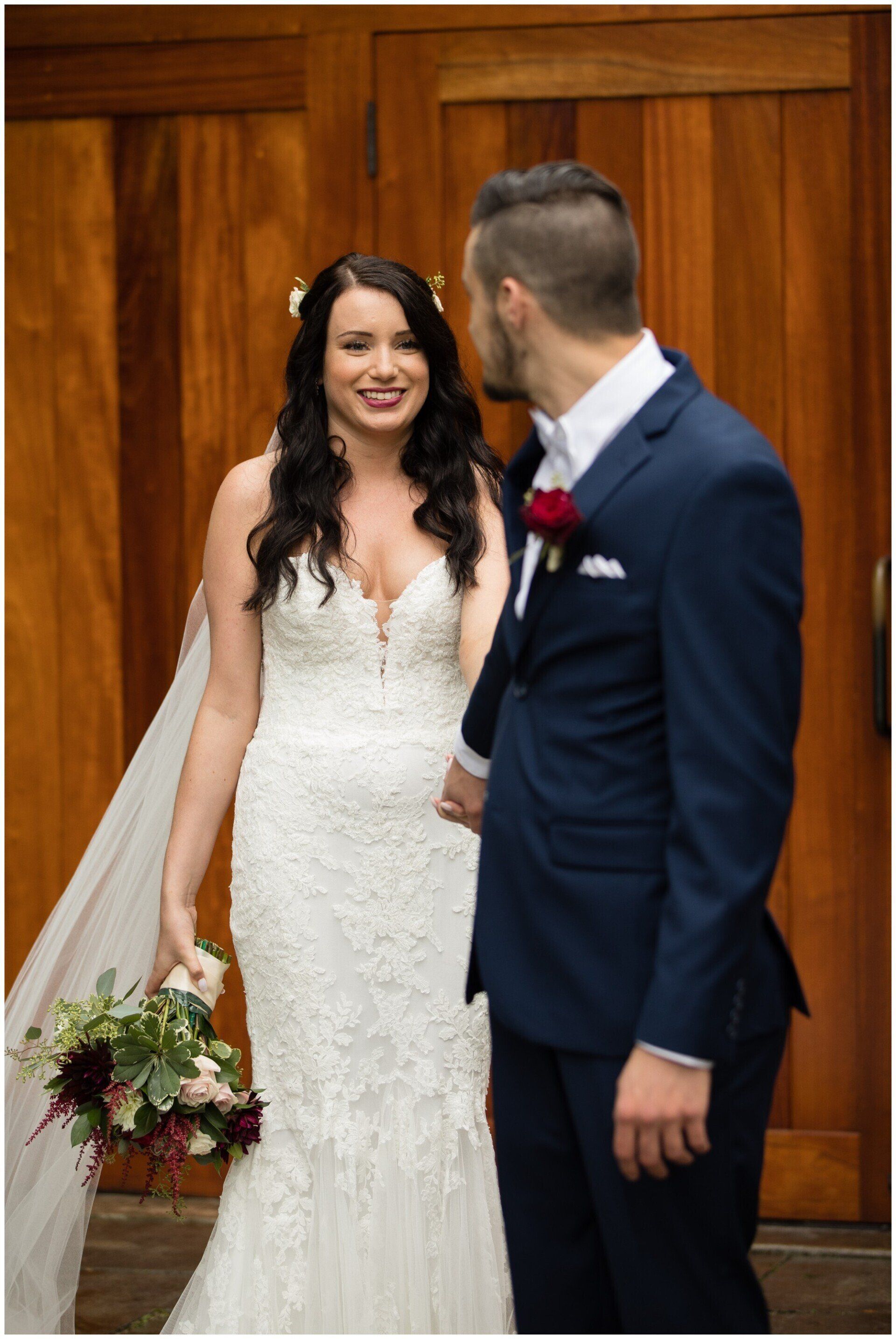 Bride in white lace gown smiling beside groom in navy suit before wooden doors