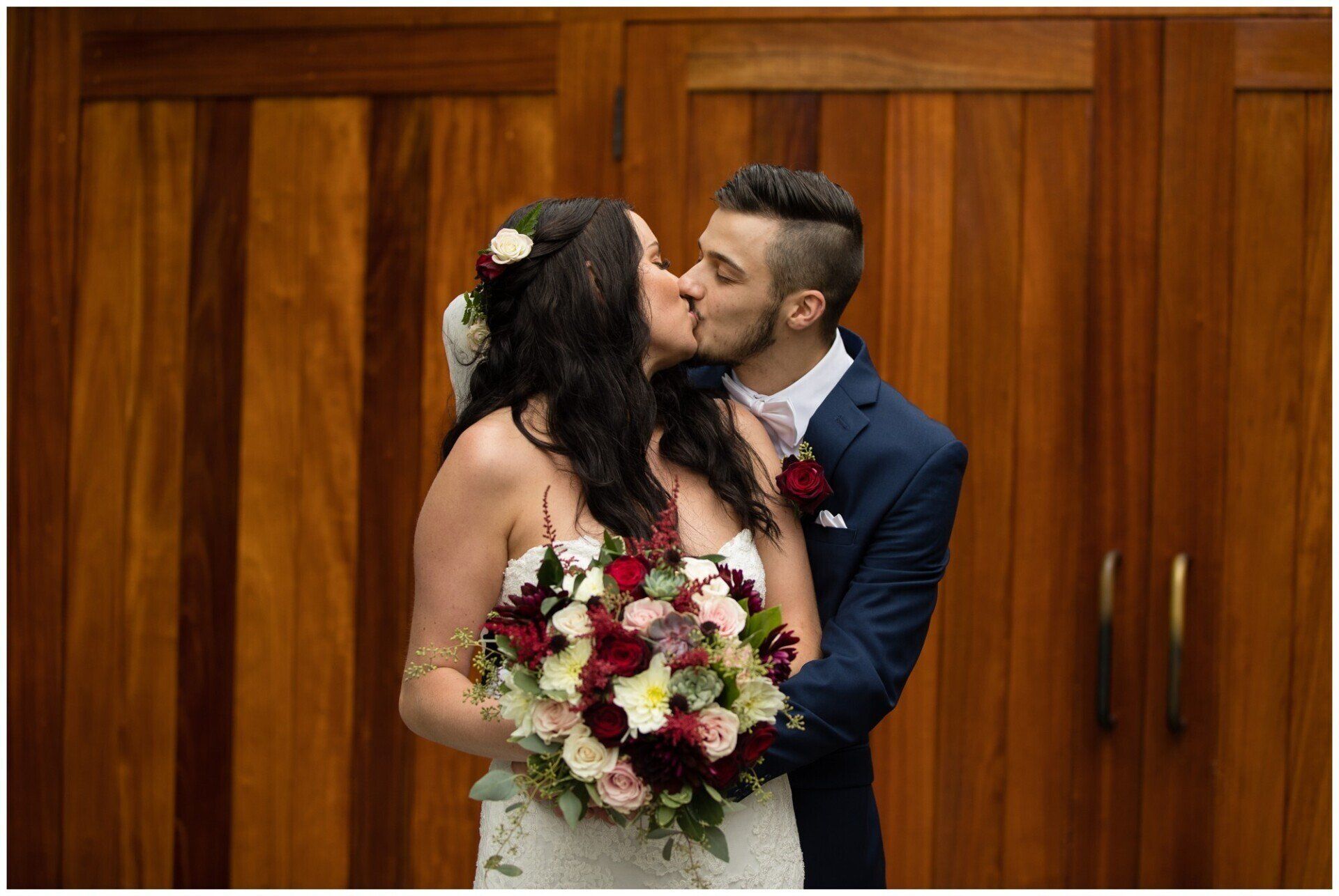 Couple kissing during wedding ceremony, bride holding a bouquet before wooden doors.
