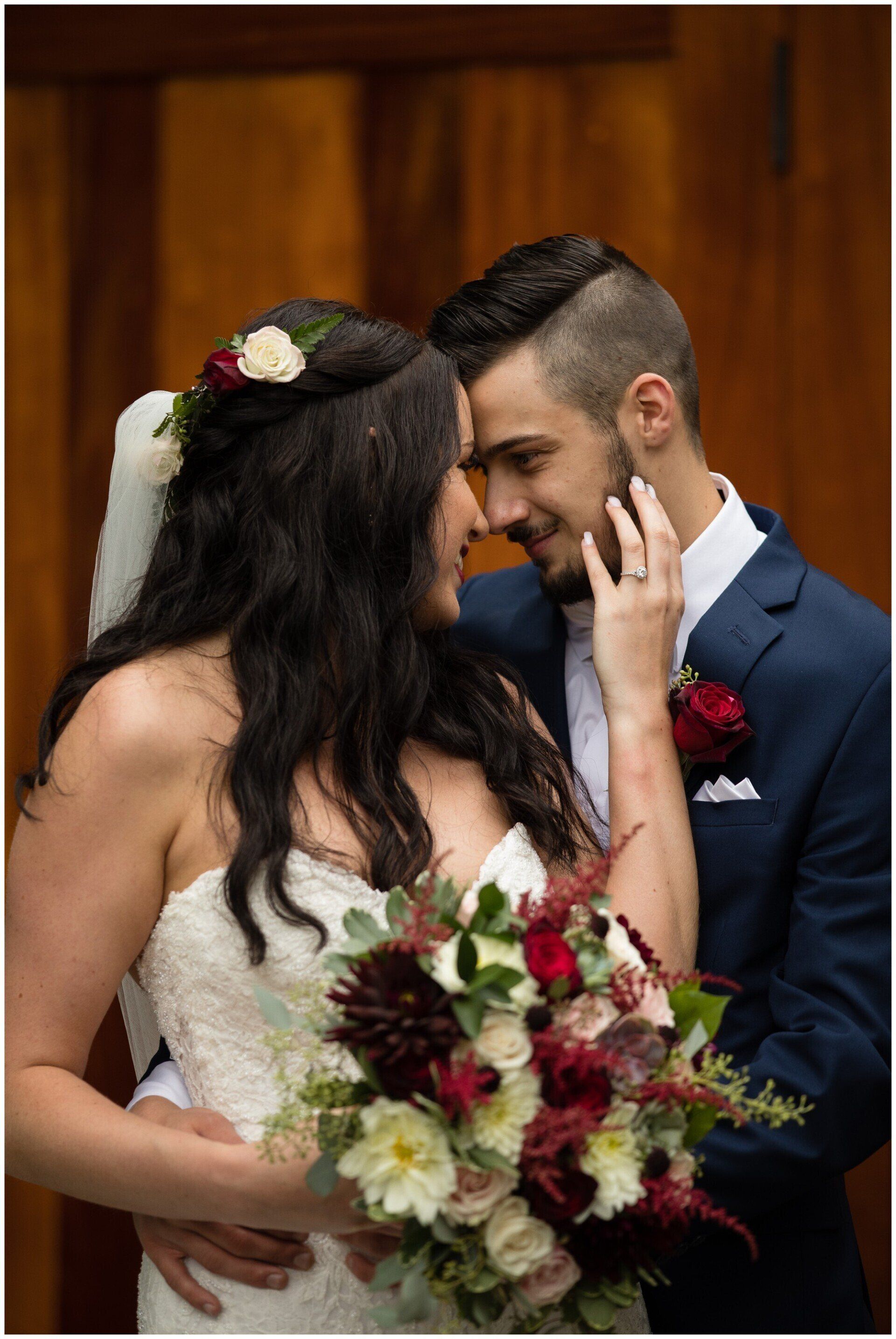 Bride and groom embrace during wedding ceremony, holding a bouquet with red and white flowers.