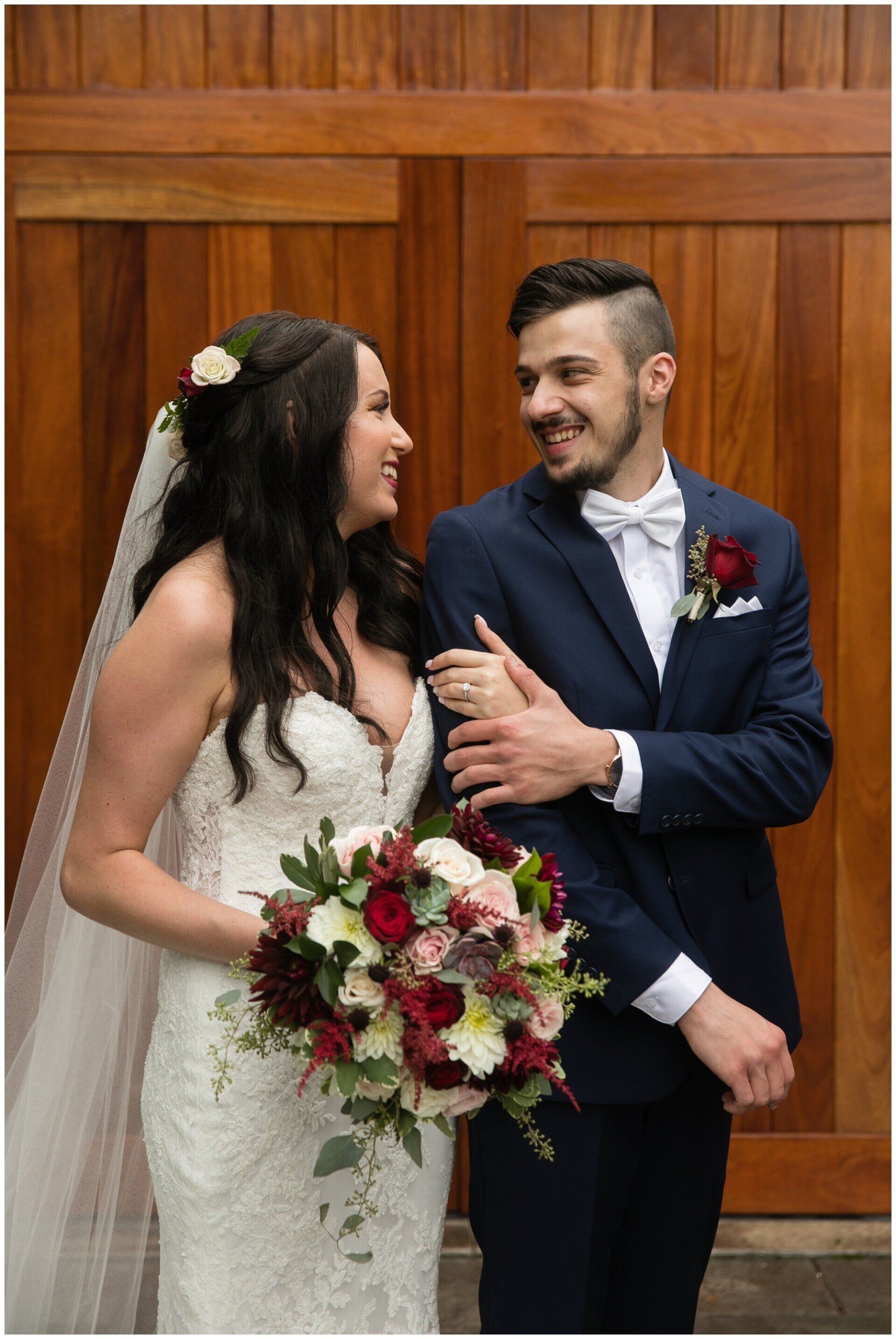 Bride and groom smiling together in wedding attire, holding a bouquet before wooden doors
