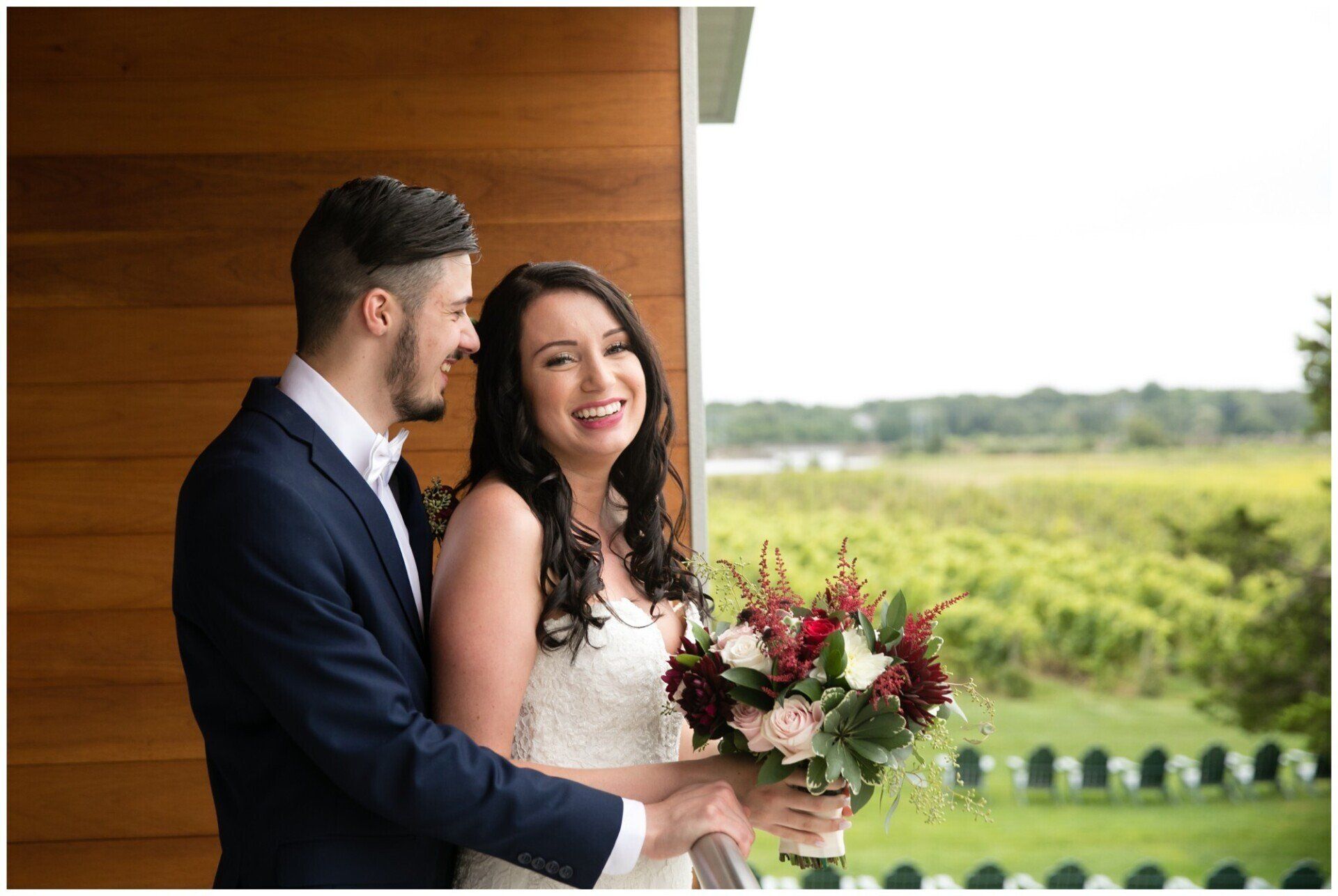 Couple in wedding attire smiling on a balcony with a bouquet, overlooking a green landscape.
